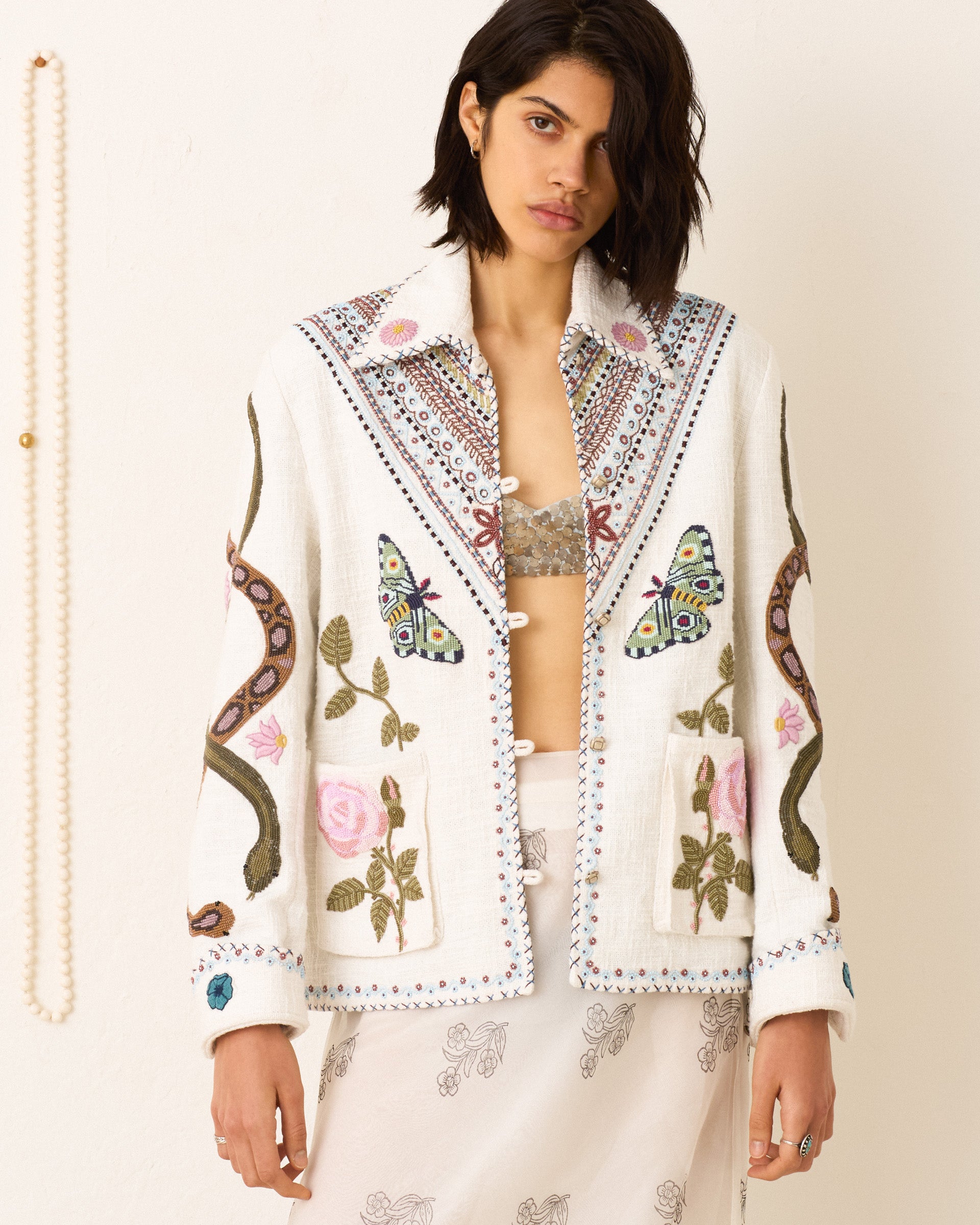 A person with dark hair wears the Jude Ivory Ethereal Jacket, featuring vintage-inspired embroidery of butterflies, snakes, and florals over a sheer patterned skirt, standing against a plain light background.