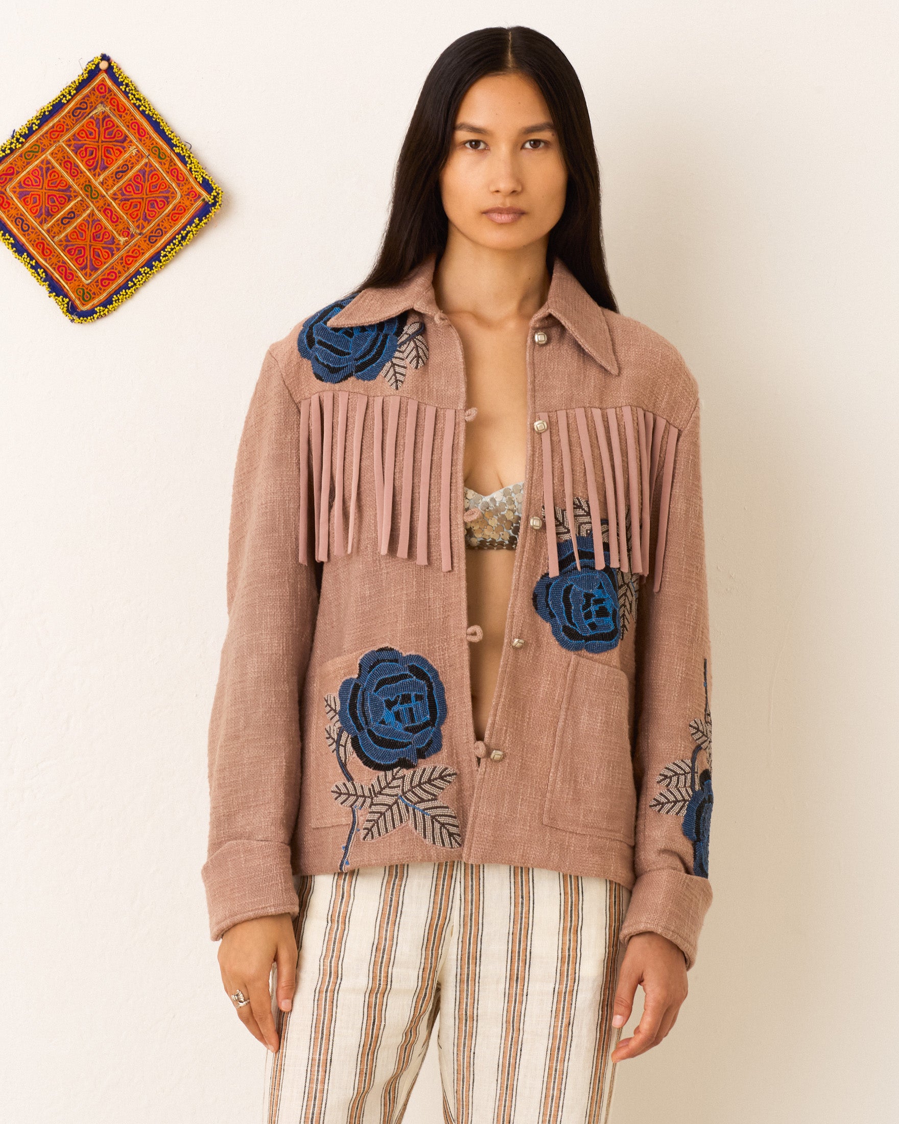 A woman stands against a white wall wearing the Jude Midnight Rosebud Jacket with fringe and blue floral embroidery, striped pants, and a patterned textile hanging beside her.