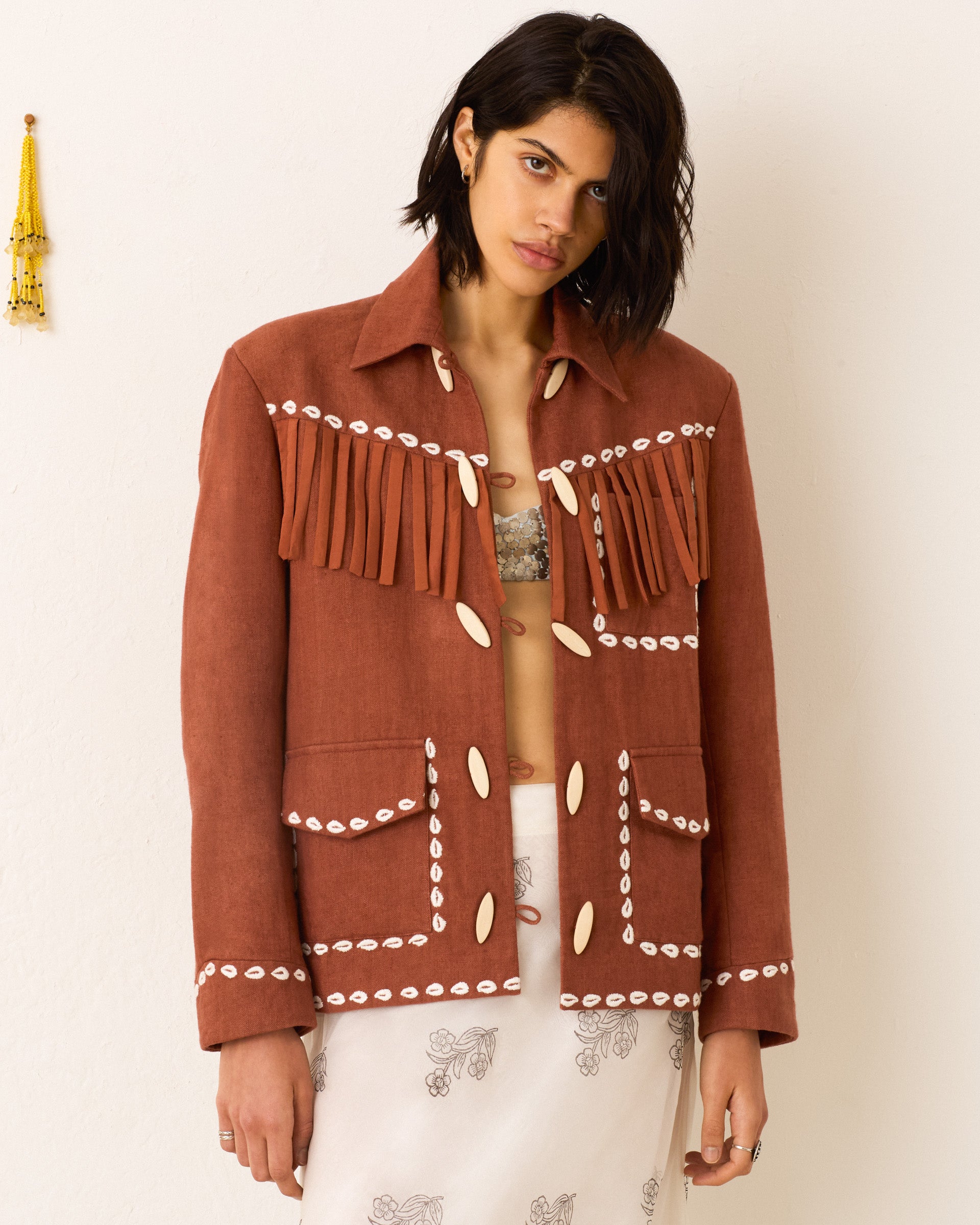 A woman wears the Jude Redwood Cowrie Jacket, a vintage-inspired rust piece with white decorative stitching and fringe details, standing against a plain wall with a tassel visible on the left.