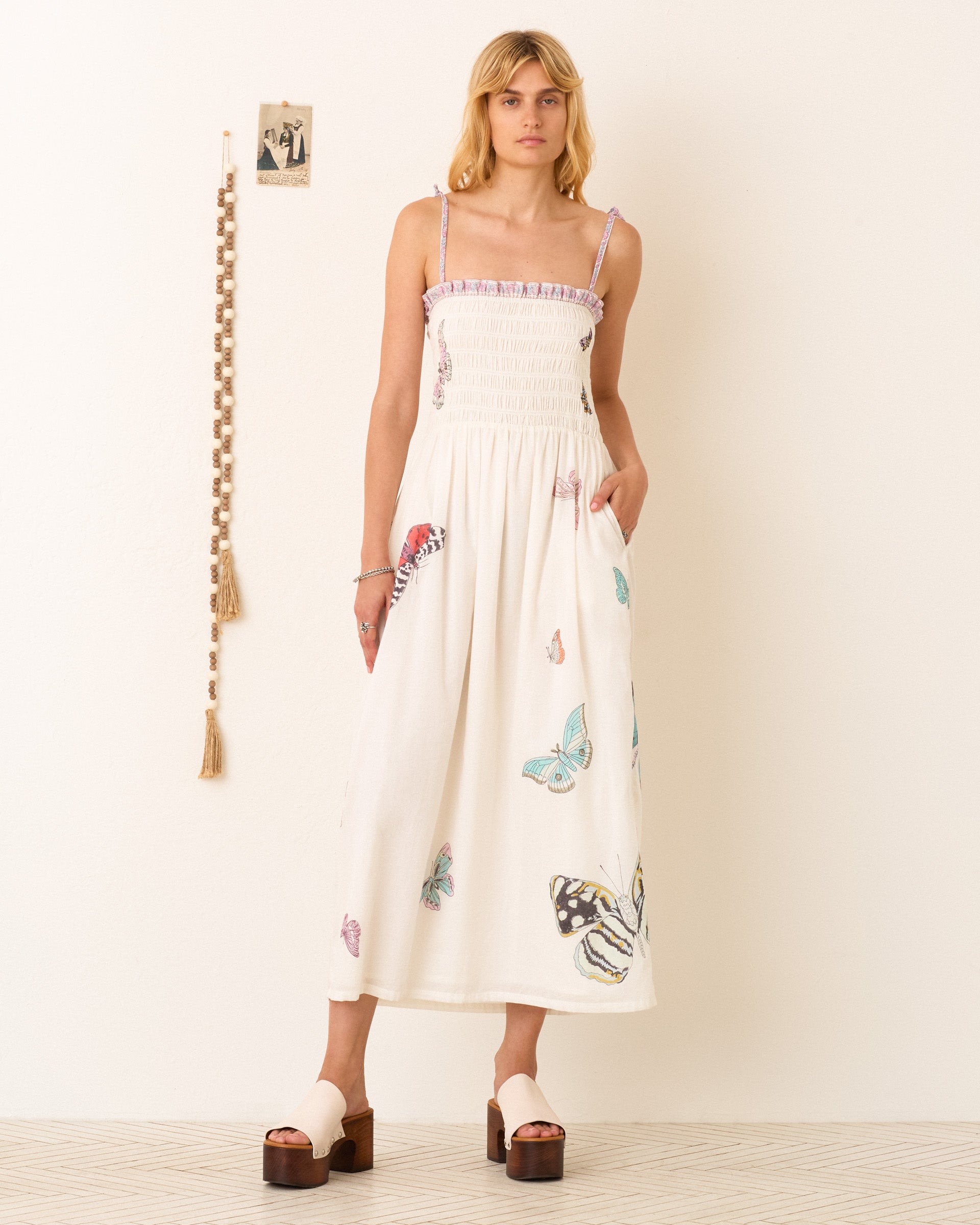 A woman stands indoors against a light wall, wearing the Kaia Butterfly Garden Dress—a hand-blocked, white ankle-length dress with butterfly prints—paired with platform sandals and minimal jewelry.