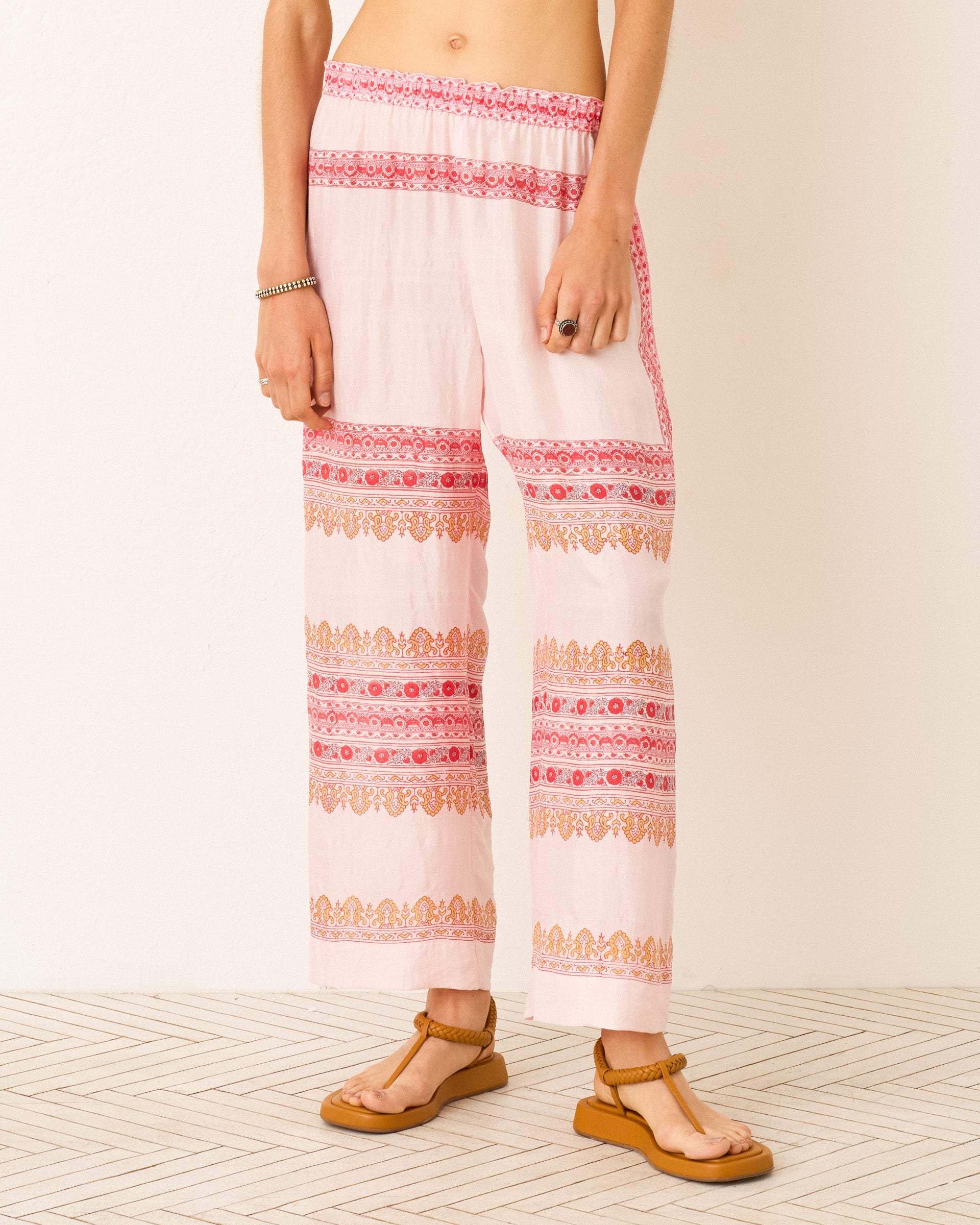 A person wears the Otto Blush Tile Pant—hand-loomed silk in light pink with red and orange horizontal stripes—paired with brown sandals and a bracelet, standing against a cream wall.
