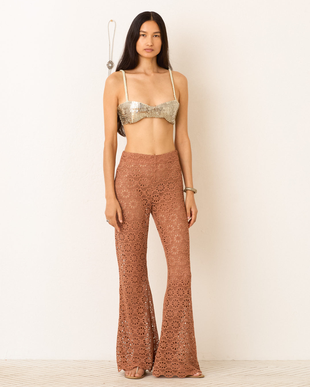 A woman stands against a plain wall wearing a metallic bralette top and the Farah Mocha Daisy Pant, high-waisted brown cotton lace bell-bottom flares, with a bracelet on her wrist.