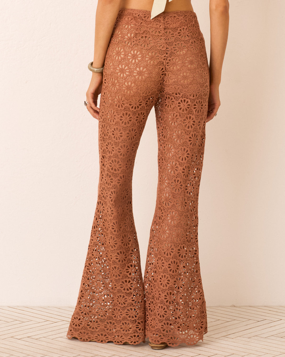 Model stands with her back to the camera against a light background, wearing the Farah Mocha Daisy Pant—high-waisted brown cotton pants with lace detail and a bell-bottom flare.