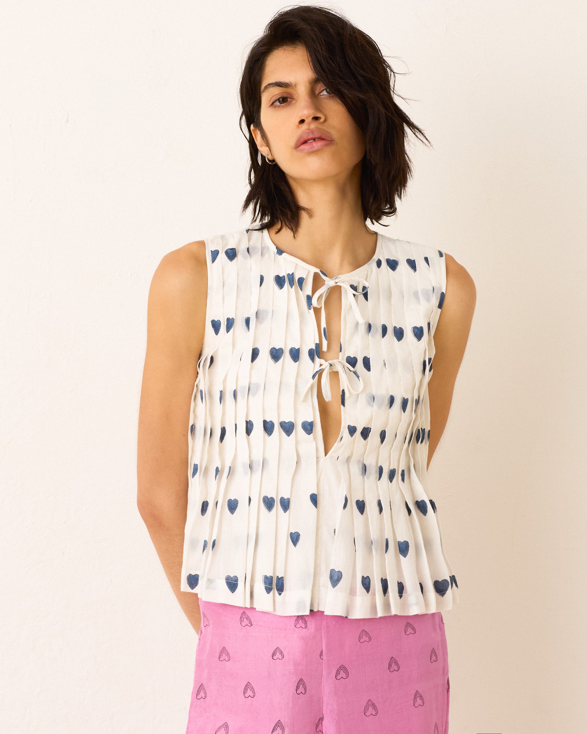 A woman wears the Rhoe Indigo Heart Top, a sleeveless hand-block printed white tie-front top with blue heart motifs, paired with high-waisted pink patterned pants, standing against a plain backdrop.