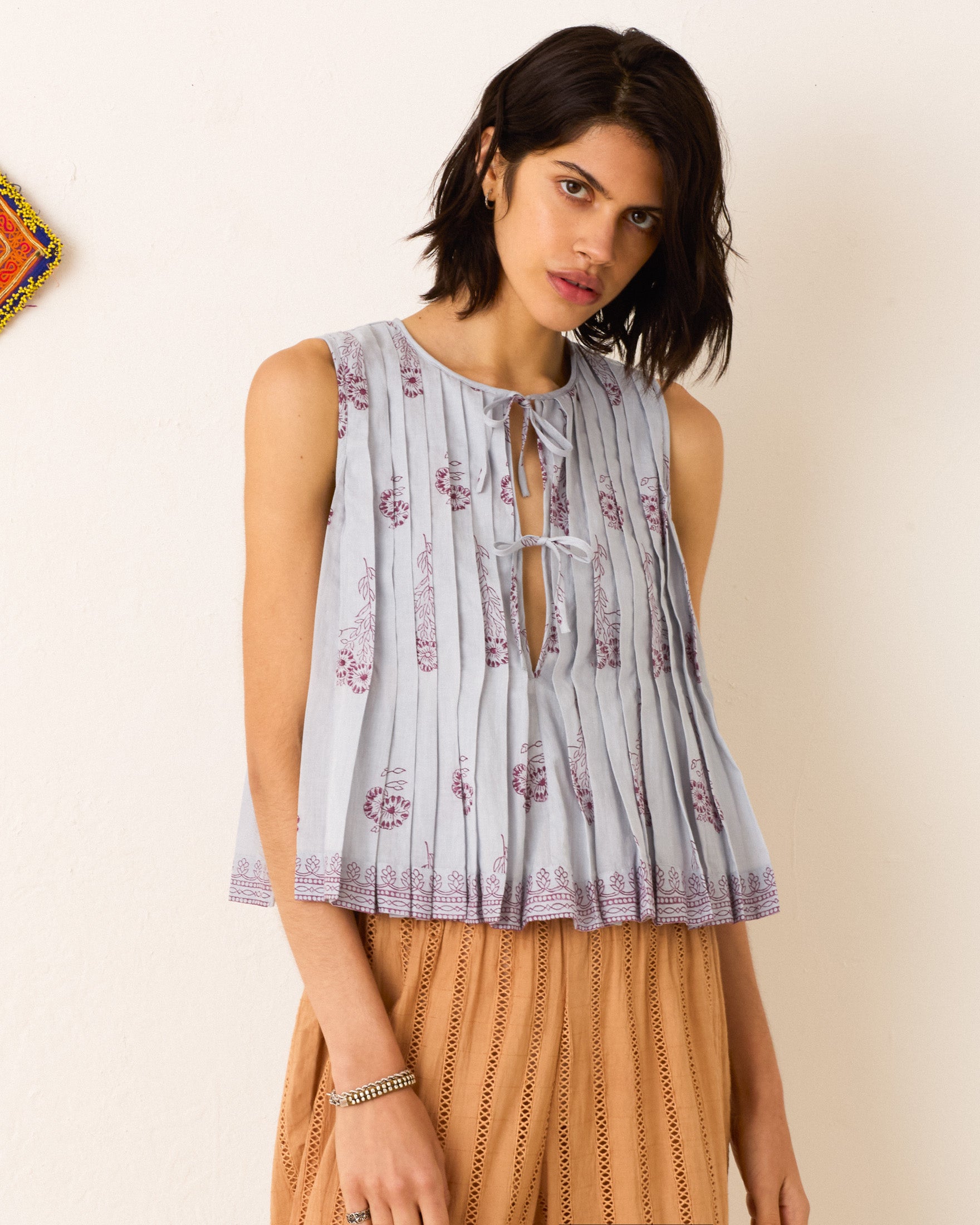 A woman wears the Rhoe Pansy Sky Top—a sleeveless light blue cotton voile top with a tie detail and maroon hand-block prints—paired with a light brown skirt, standing against a plain white wall.