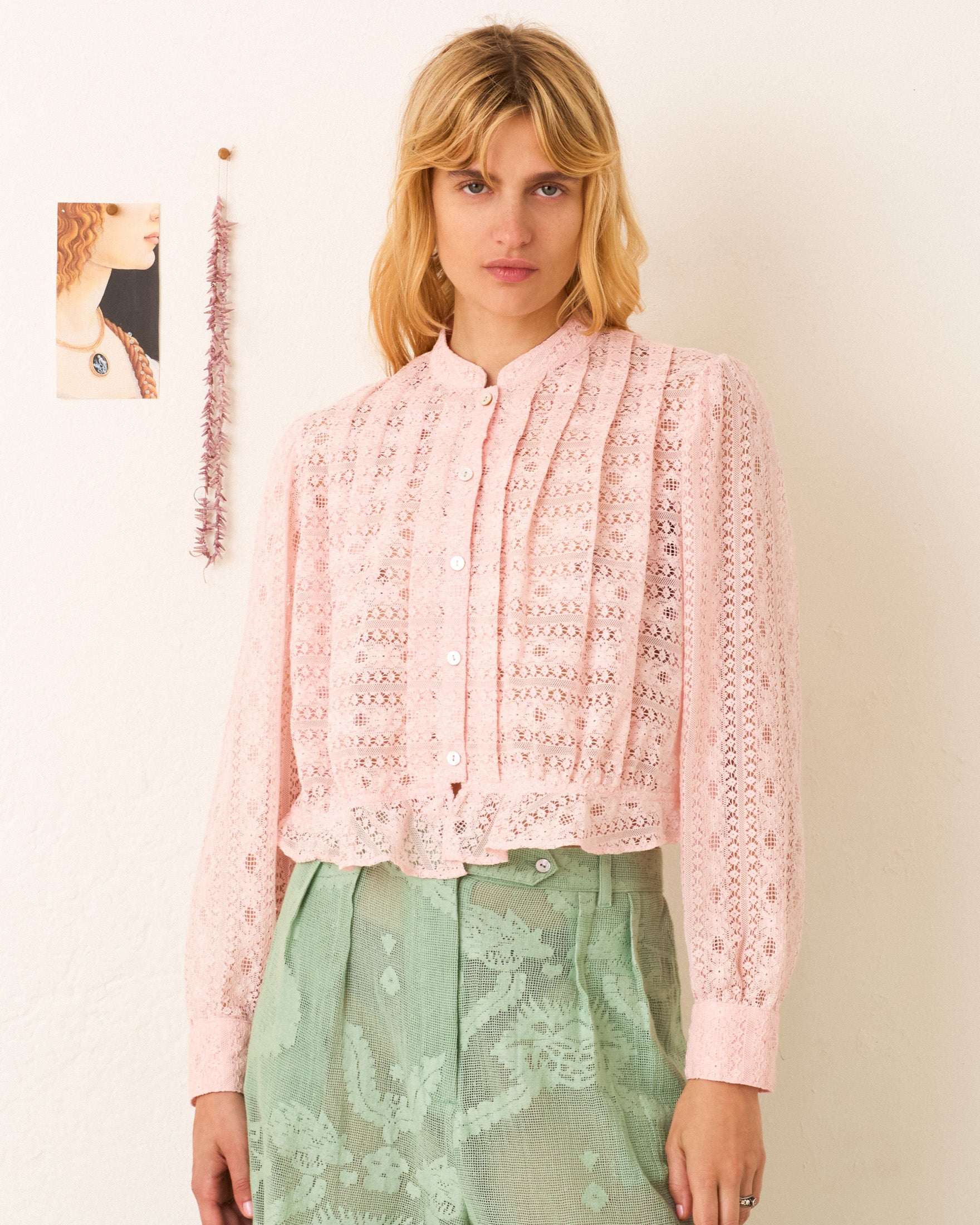 A woman wearing the Ryland Blush Lace Shirt and pale green high-waisted trousers stands against a white wall decorated with a small photo and hanging string.