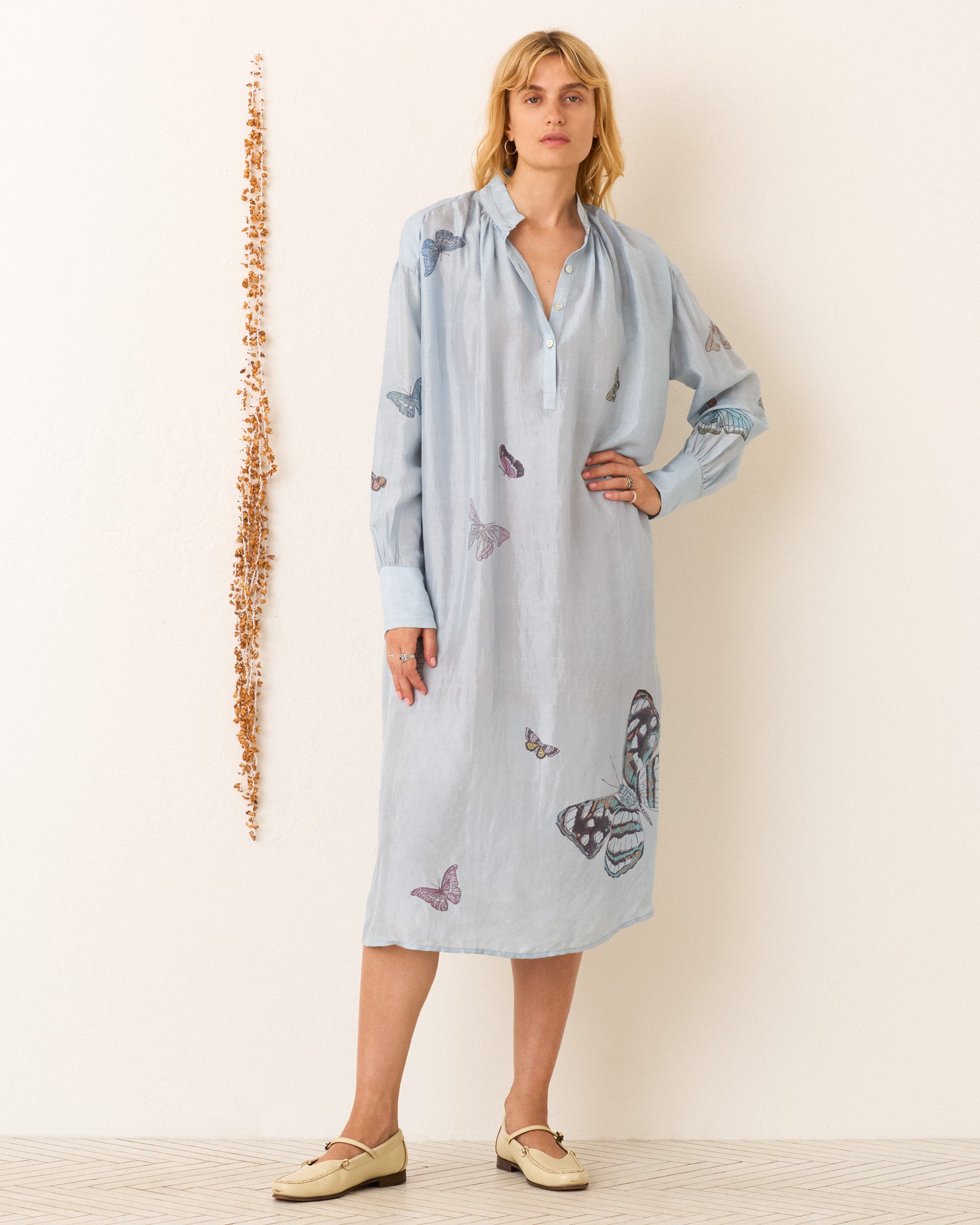 A woman stands against a white wall wearing the Shawna Powder Butterfly Dress—light blue, hand-block printed, long-sleeved with butterfly prints—and beige loafers. A vertical string of shells hangs beside her.