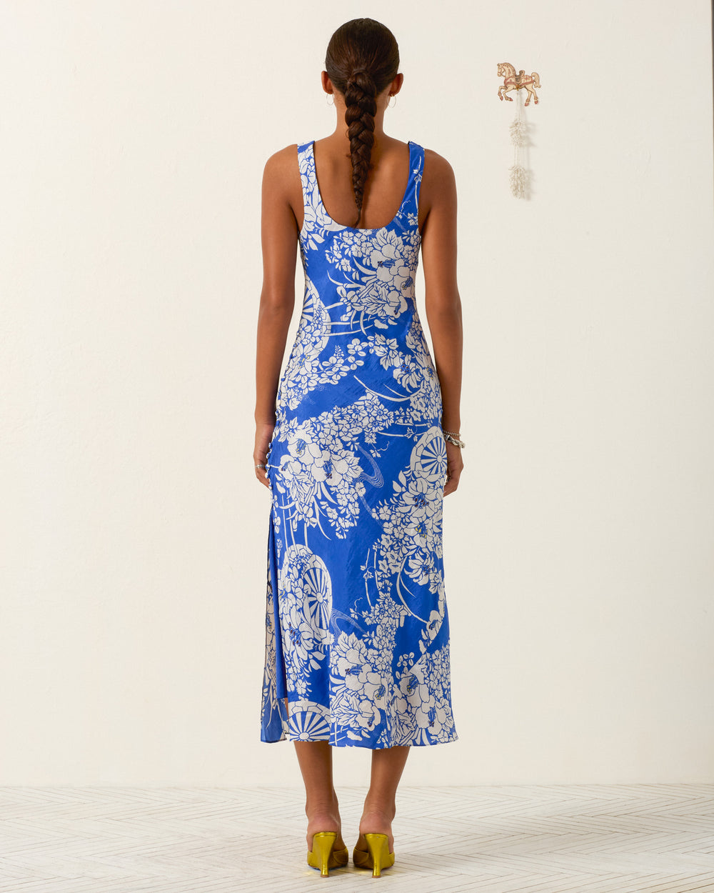 A woman with a long braid faces away, wearing the sleeveless blue and white Adele Royal Spoke Silk Dress and yellow shoes against a plain light background.