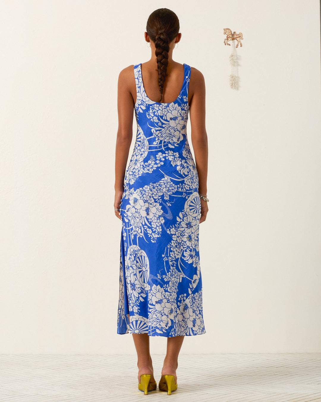 A woman with a long braid faces away, wearing the sleeveless blue and white Adele Royal Spoke Silk Dress and yellow shoes against a plain light background.