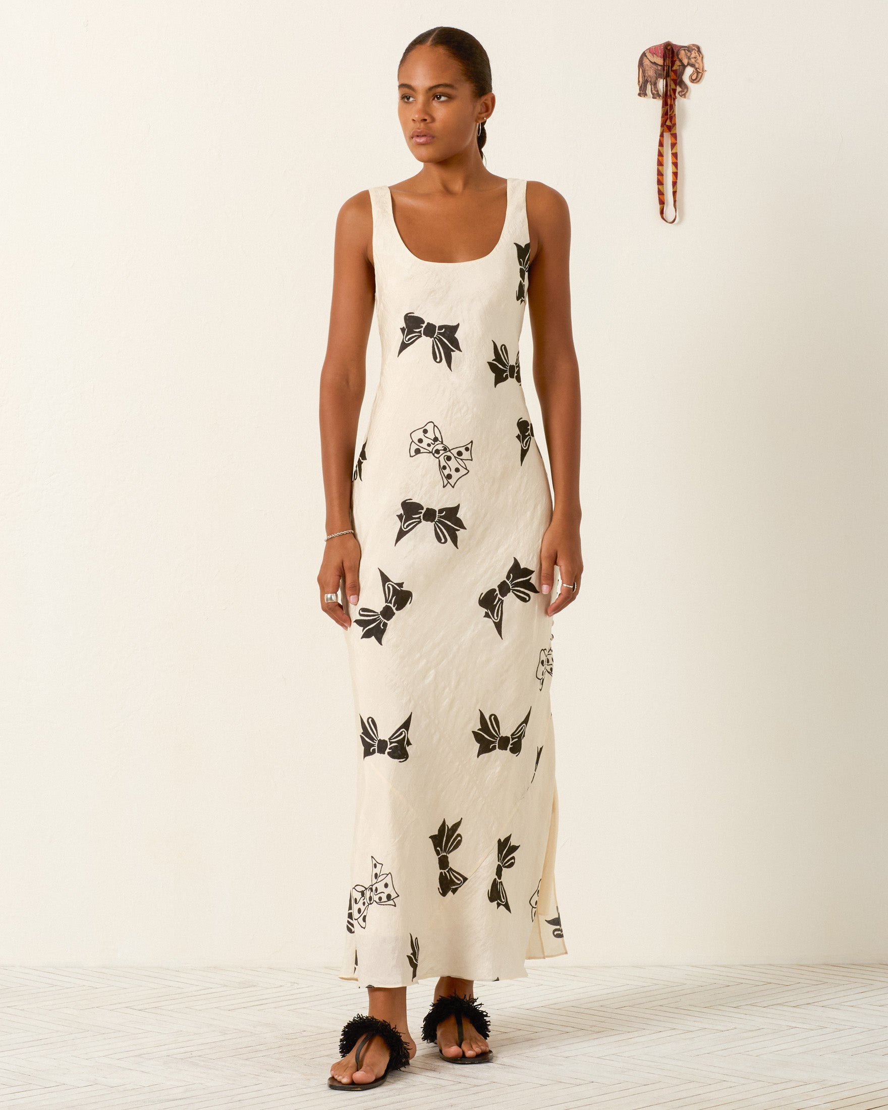 A woman stands indoors wearing the Adele Black Tie Silk Dress, a sleeveless, ankle-length cream dress adorned with black floral hand-block prints and styled with black sandals; decorative items hanging on the wall in the background accentuate the artisanal charm of this unique garment.