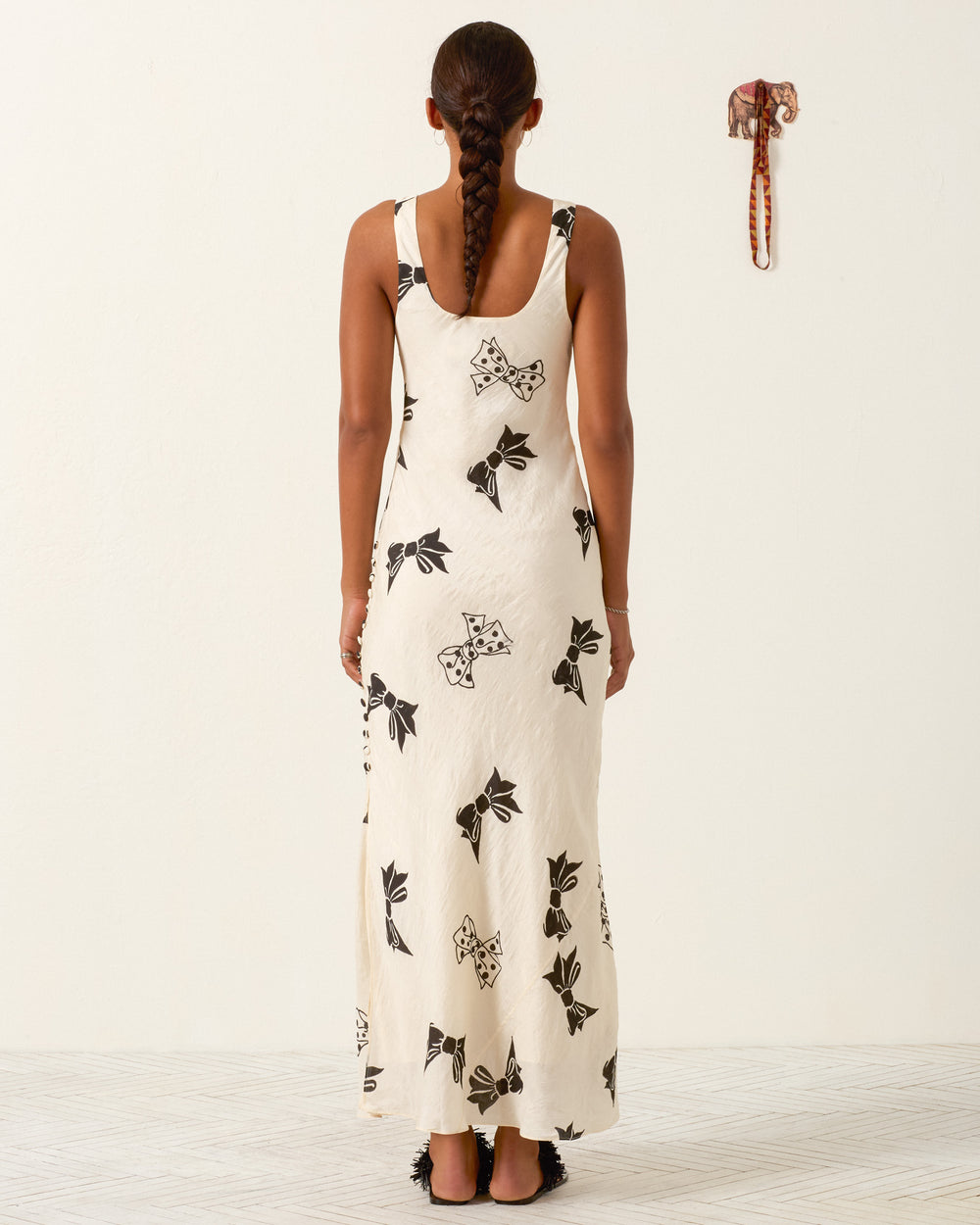 A woman with a braid stands facing away, wearing the Adele Black Tie Silk Dress—a long white hand-block-printed dress featuring black floral patterns; a small elephant wall decoration is visible on a plain white wall.