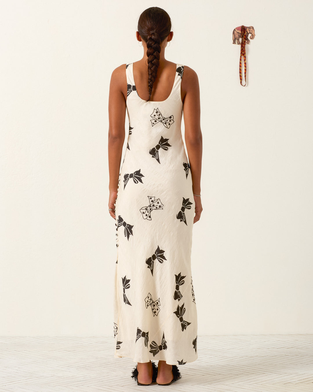 A woman with a braid stands facing away, wearing the Adele Black Tie Silk Dress—a long white hand-block-printed dress featuring black floral patterns; a small elephant wall decoration is visible on a plain white wall.