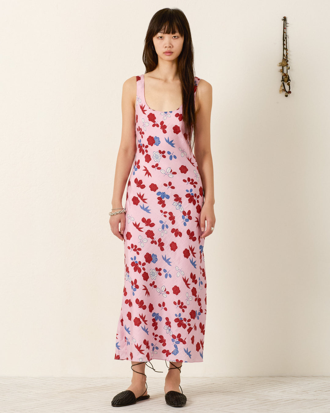 A woman stands indoors wearing the Adele Tutu Bloom Silk Dress, a sleeveless, ankle-length pink slip dress with a red and blue floral pattern, paired with black flats and bracelets.