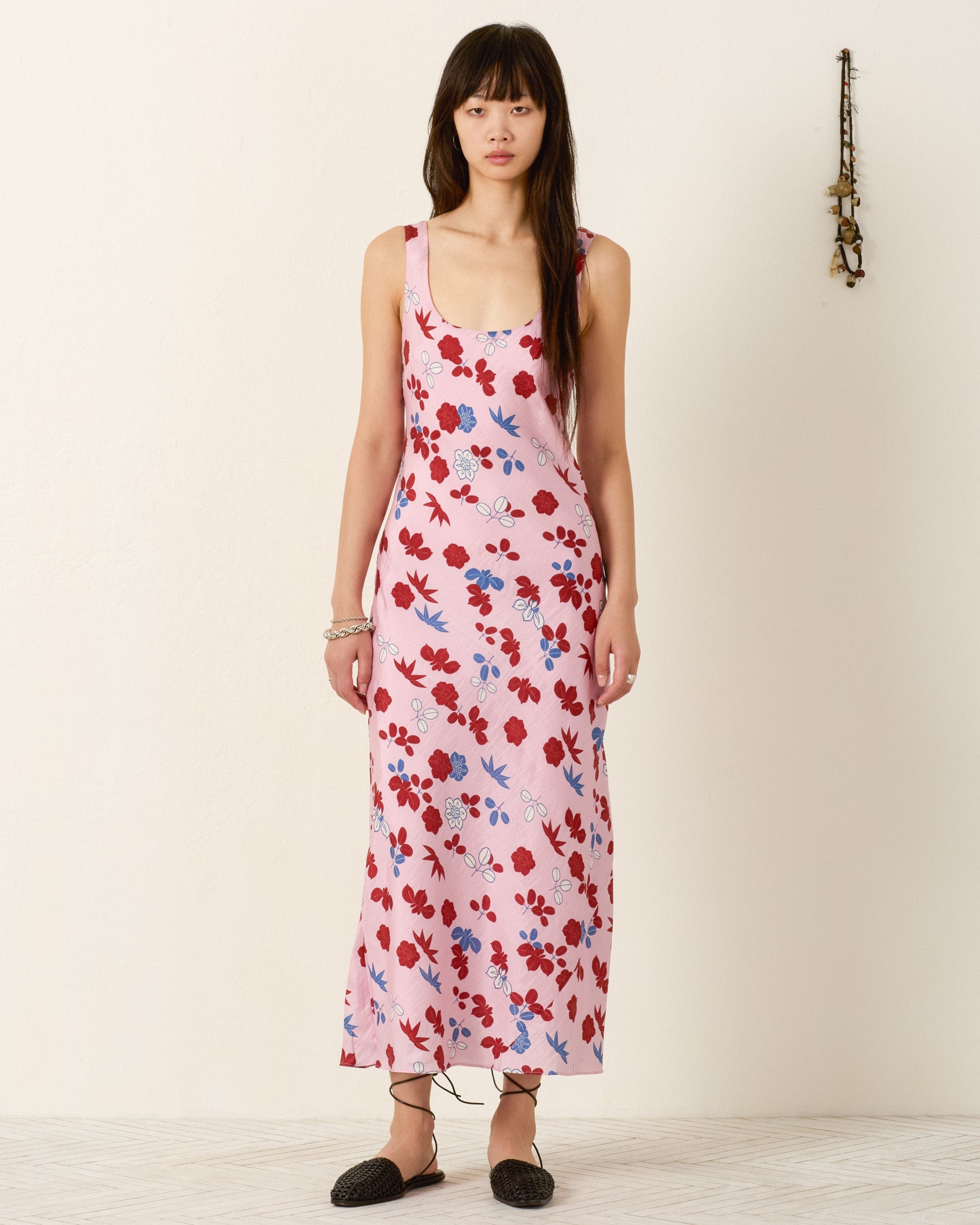 A woman stands indoors wearing the Adele Tutu Bloom Silk Dress, a sleeveless, ankle-length pink slip dress with a red and blue floral pattern, paired with black flats and bracelets.