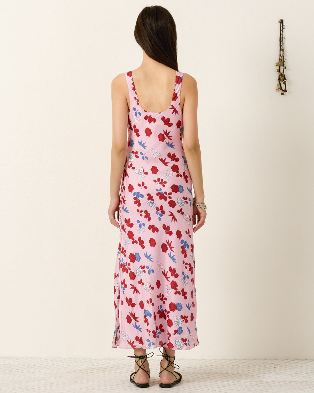 A woman stands with her back to the camera in the Adele Tutu Bloom Silk Dress, an ankle-length, bias-cut slip dress in a red, white, and blue floral pattern that drapes elegantly for a modern silhouette. She pairs it with black sandals.