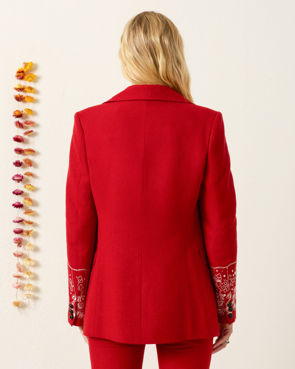 A person with long blonde hair, seen from behind, wears the Afternoon Russet Lace Blazer with notched lapel and embroidered cuffs, standing near a wall decorated with a vertical line of dried flowers.