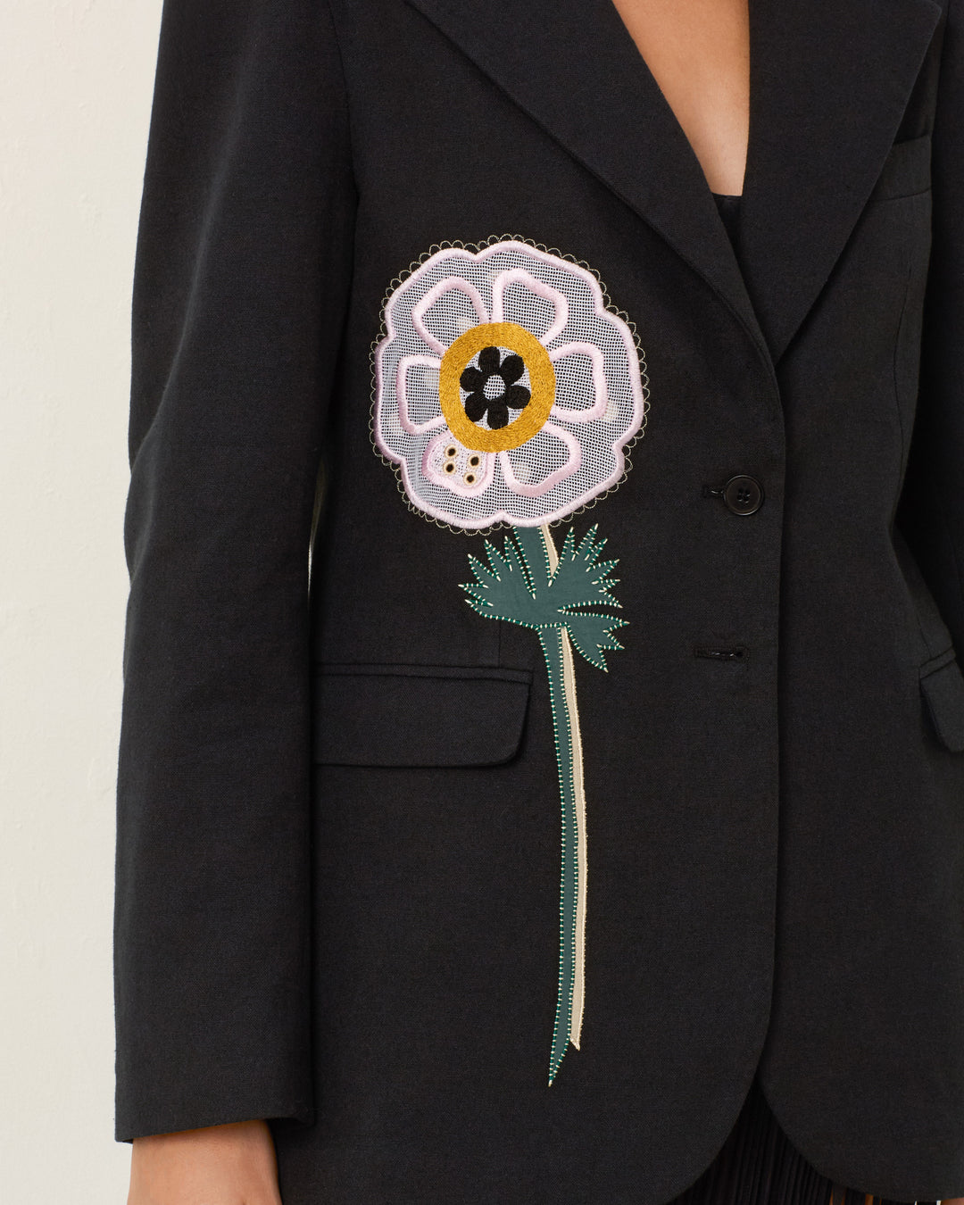 A person wearing the Afternoon Pressed Daisy Blazer, a black blazer featuring a large handmade daisy embroidery on one side.