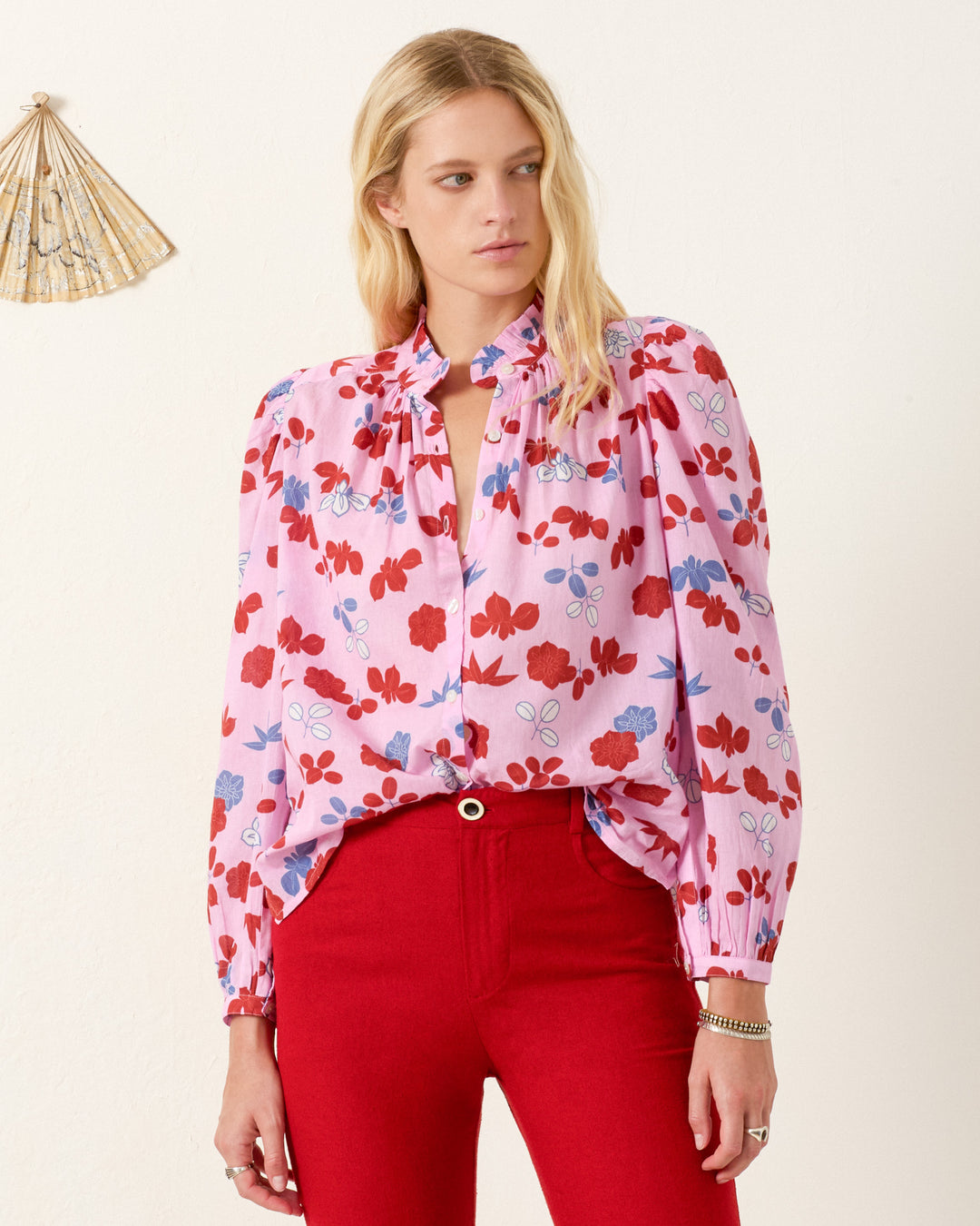 A woman with long blonde hair wears the Annabel Tutu Bloom Shirt, a pink floral cotton voile top with puff shoulders, tucked into high-waisted red pants. She stands against a plain background with a decorative fan on the wall.