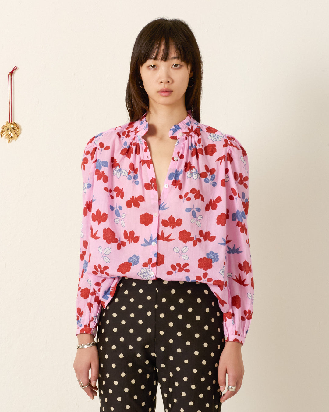 A woman stands against a plain wall wearing the Annabel Tutu Bloom Shirt in pink cotton voile with puff shoulders and black polka dot pants. A small ornament hangs on the wall beside her.