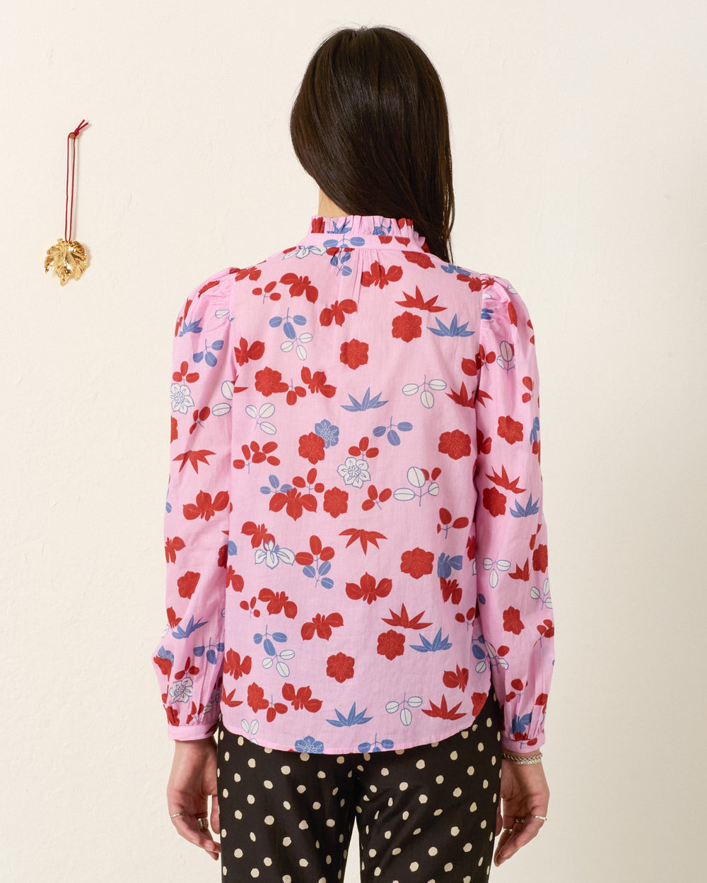 A woman with long dark hair faces a wall, wearing the Annabel Tutu Bloom Shirt—a pink cotton voile top with botanical motifs—and black pants with white polka dots. A small gold ornament hangs on the wall to her left.