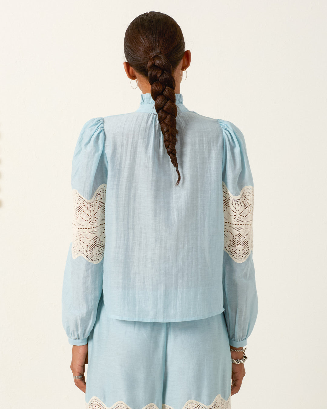 A woman with a long braid stands facing away, wearing the Annabel Sky Prima Lace Shirt—light blue cotton-silk with puff shoulders and white lace details—paired with matching shorts.