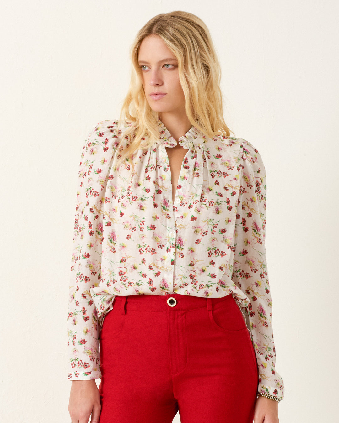 A woman stands against a plain light background, wearing the Artemis Wild Garden Shirt—a white floral blouse with a ruffled collar and keyhole neckline—paired with red high-waisted pants.