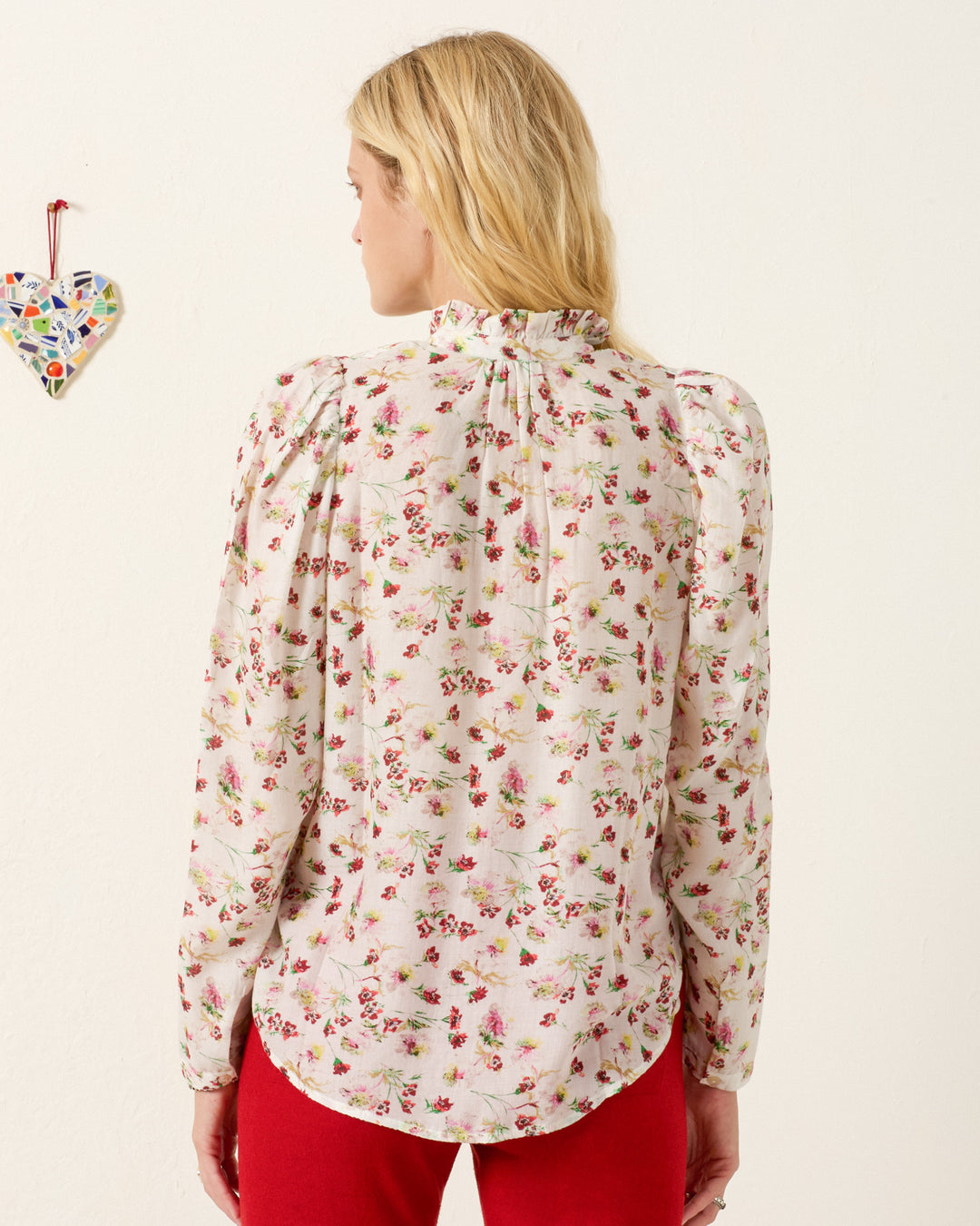 A woman with blonde hair, seen from behind, wears the Artemis Wild Garden Shirt—long-sleeved with a pink floral print and puff sleeves—paired with red pants. A small mosaic heart is on the wall.