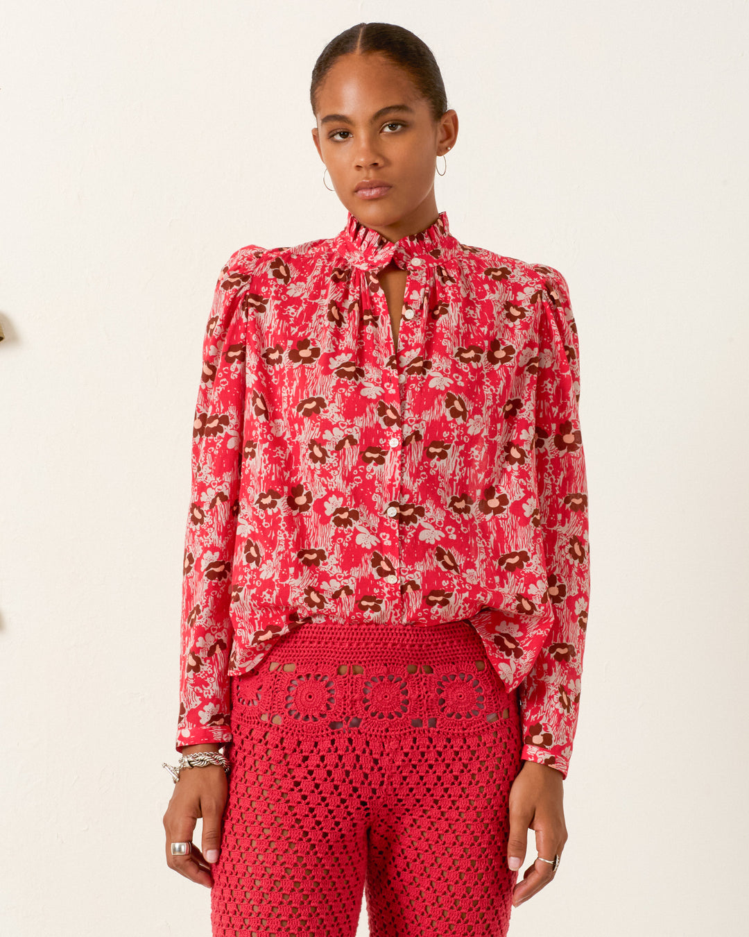 A woman faces forward, wearing the Artemis Rougewood Shirt—a red floral cotton voile piece with puff sleeves and a keyhole neckline—paired with red crocheted pants against a plain light background.