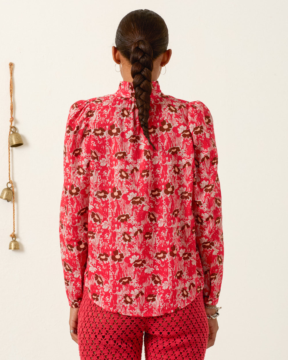 A person with a braid wears the Artemis Rougewood Shirt, a red patterned cotton voile top with puff sleeves, paired with matching pants, and stands facing a white wall decorated with hanging bells.