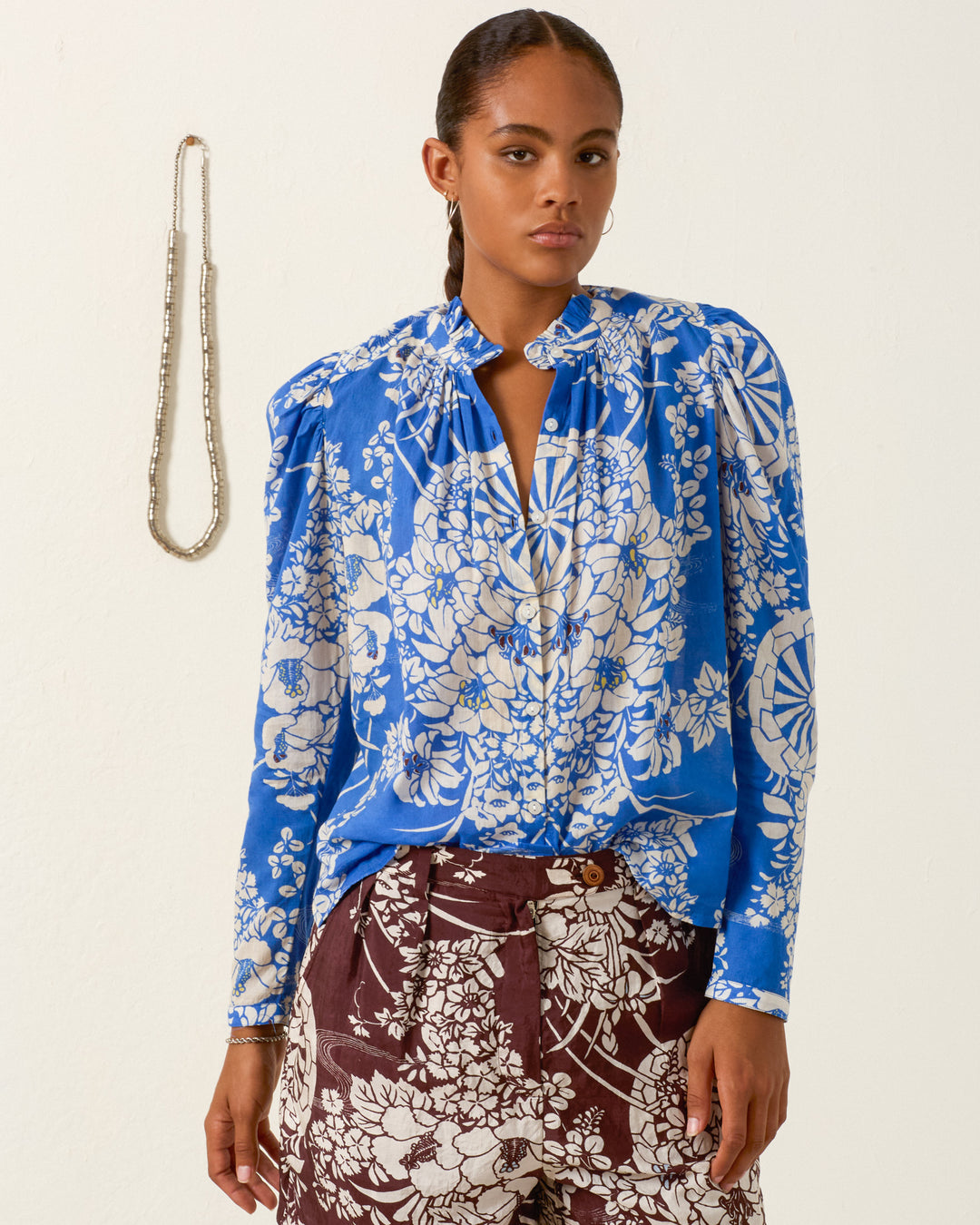 A woman stands against a white wall wearing the Artemis Royal Spoke Shirt, a blue and white cotton voile top with a ruffled collar, paired with brown floral pants; a beaded necklace hangs on the wall behind her.