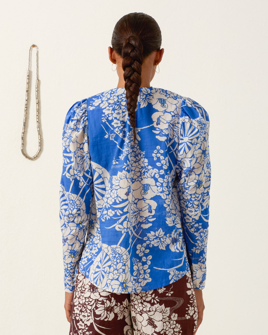 A woman with a long braid faces away, wearing the Artemis Royal Spoke Shirt—blue and white cotton voile with a ruffled collar—and brown patterned pants. A beaded necklace hangs on the white wall to her left.
