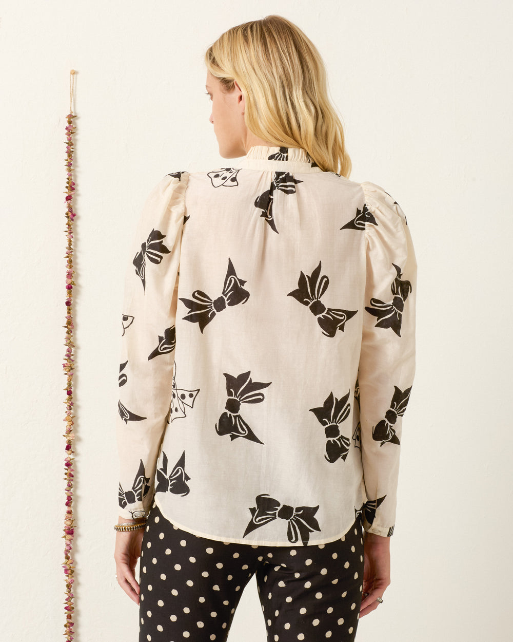 A woman stands facing away, wearing the Artemis Black Tie Shirt with a ruffled collar and black polka-dot pants, against a plain wall with a hanging decoration.