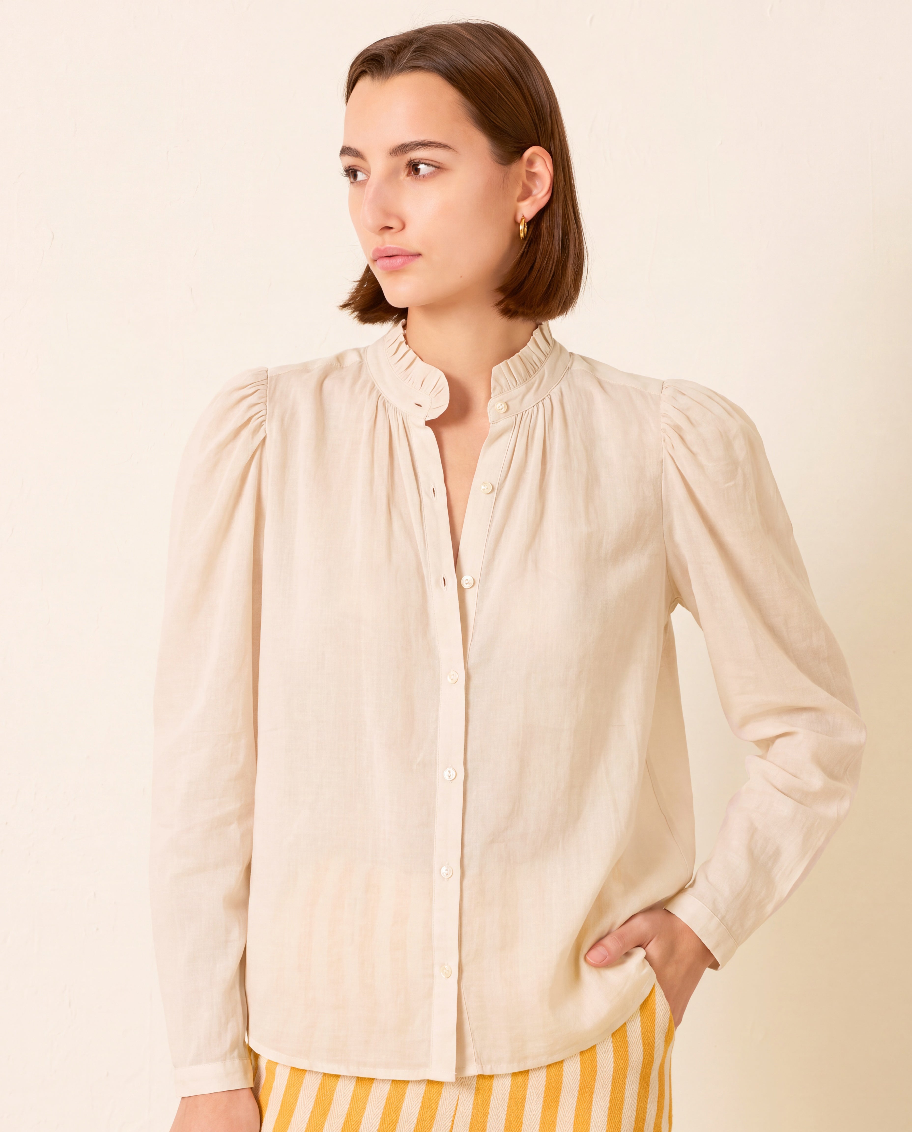 A person with straight brown hair wears the Artemis Stone Shirt—a light beige cotton voile top with a ruffled collar and buttons—standing against a plain off-white background, one hand resting on their hip.