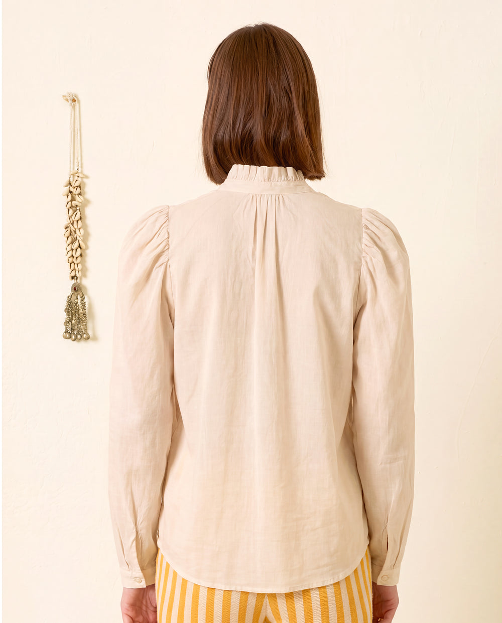 A person with short brown hair, seen from behind, wearing the Artemis Stone Shirt—a long-sleeved cream cotton voile shirt with a delicate ruffled collar—and yellow striped pants, stands against a plain wall with a hanging decoration.