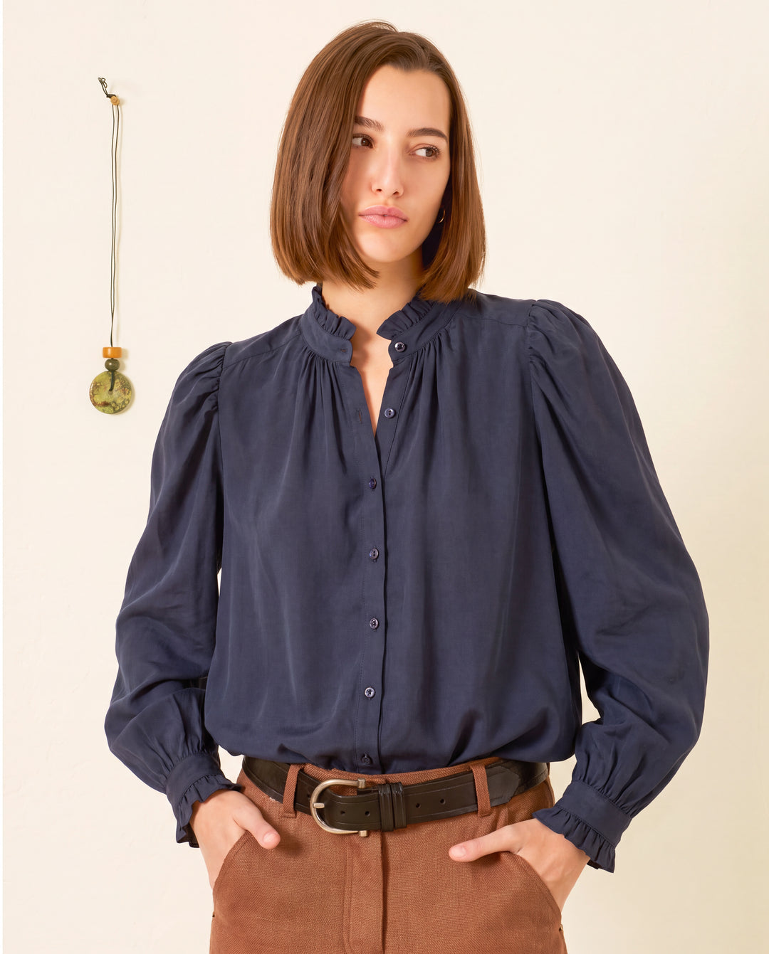 A woman wears the Artemis Deep Sea Shirt—a navy cotton voile top with puff sleeves—paired with brown pants, standing against a light wall adorned with a small hanging ornament.