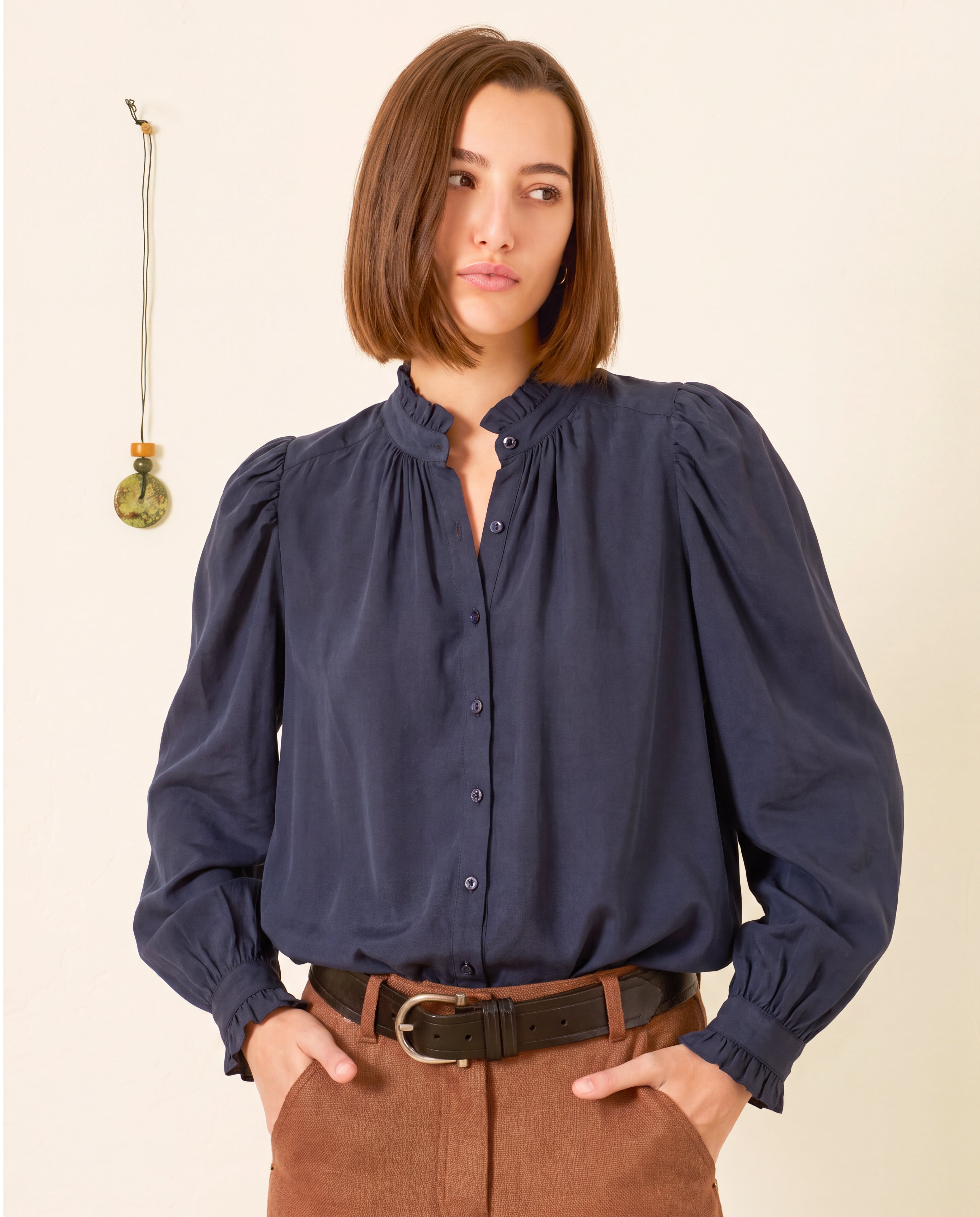 A woman wears the Artemis Deep Sea Shirt—a navy cotton voile top with puff sleeves—paired with brown pants, standing against a light wall adorned with a small hanging ornament.