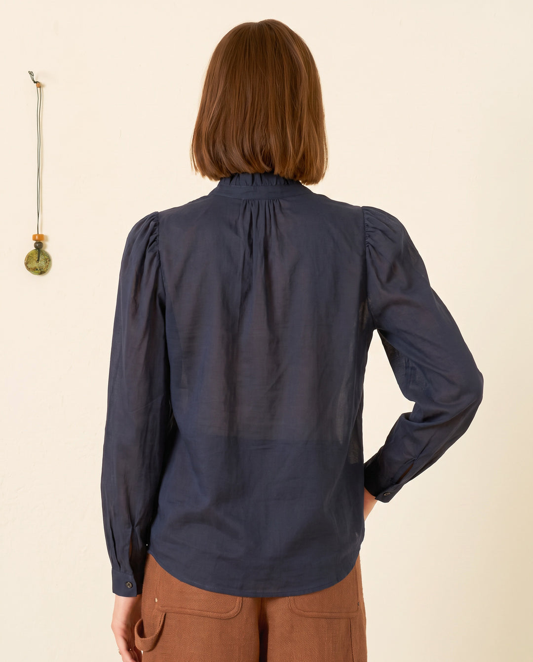A person with chin-length brown hair, shown from the back, is wearing the Artemis Deep Sea Shirt—a navy long-sleeve cotton voile top with subtle puff sleeves—paired with brown pants, standing indoors near a cream wall adorned with a small hanging ornament.