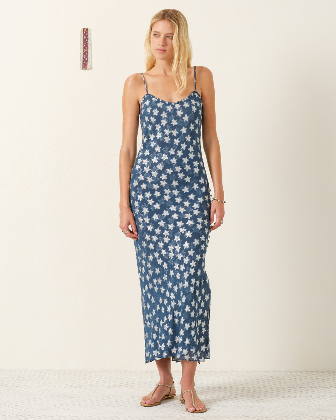 A woman stands against a plain wall wearing the Athens Starlite Dress—a blue, ankle-length viscose-chiffon slip with a white star pattern, thin straps, sweetheart neckline—and beige sandals.