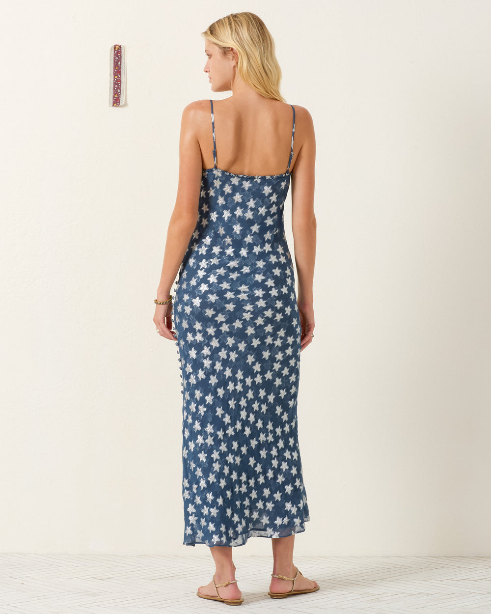 A woman stands in a room facing away, wearing the Athens Starlite Dress—a blue ankle-length viscose-chiffon slip dress with white star patterns—paired with sandals. A small patterned wall decor appears in the background.