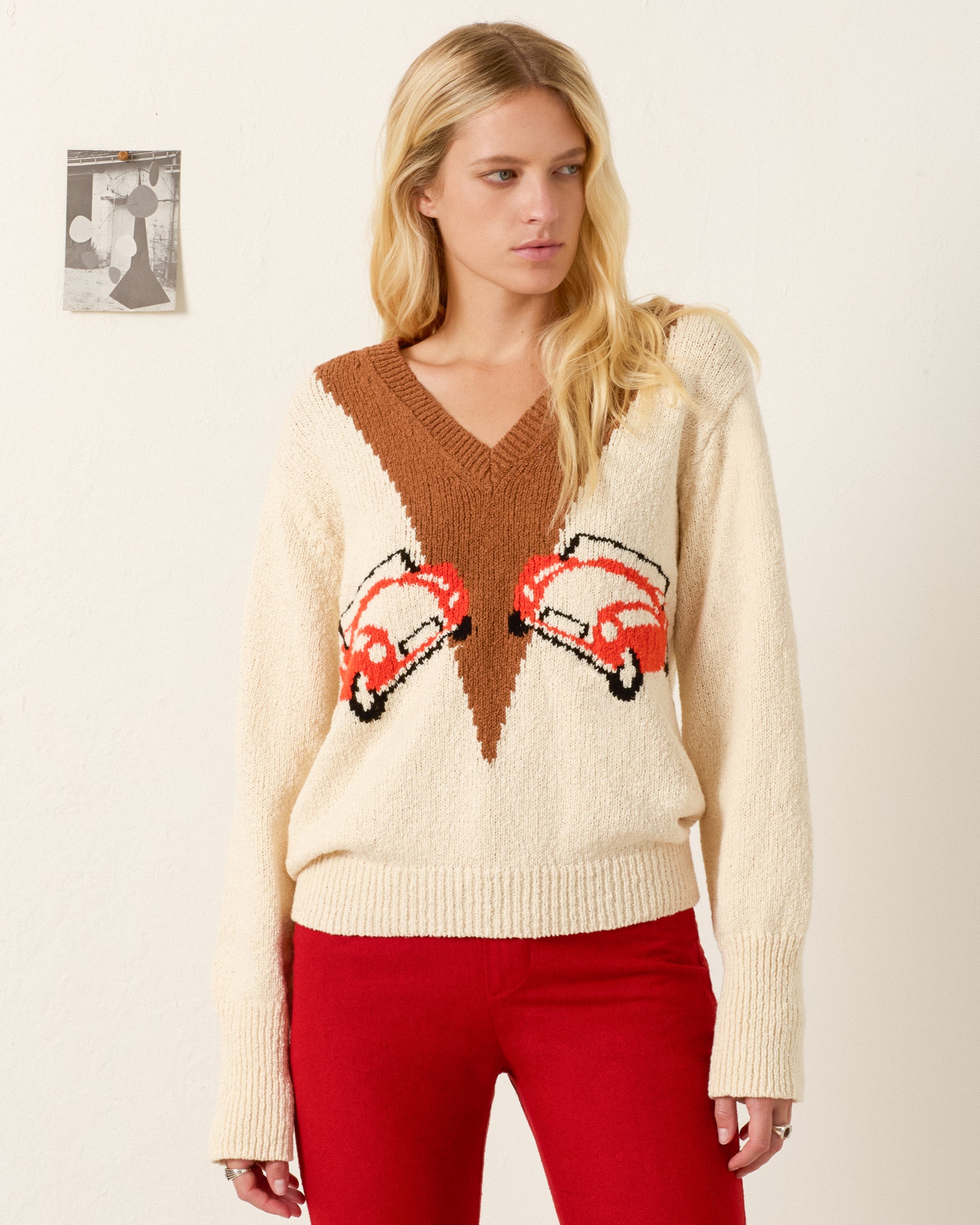 A woman wearing the Axel Vintage Cruiser Sweater with a vintage car motif and red pants stands in front of a white wall with a small photo taped to it.