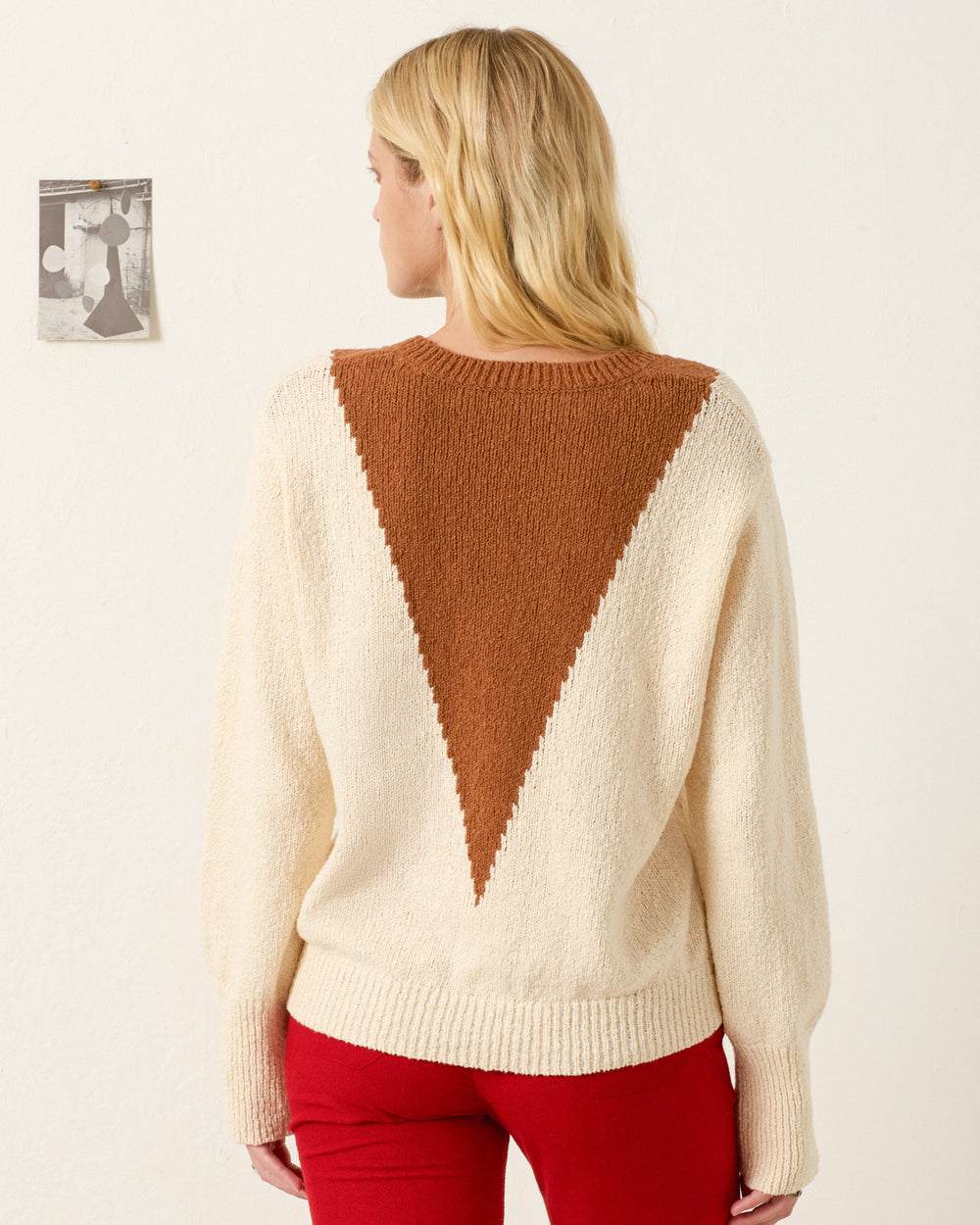 A person with long blonde hair, wearing the Axel Vintage Cruiser Sweater—an Italian cotton V-neck with a large brown triangle design on the back—and red pants, stands facing away from the camera. A small black and white photo is on the wall.