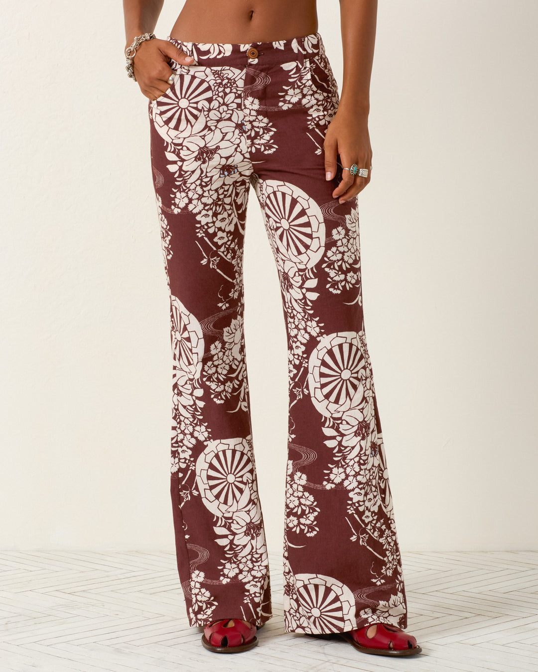 A person wearing the Bunny Auburn Spoke Jean—high-waisted, maroon flared pants with a white hanaguruma motif—paired with red open-toe sandals.