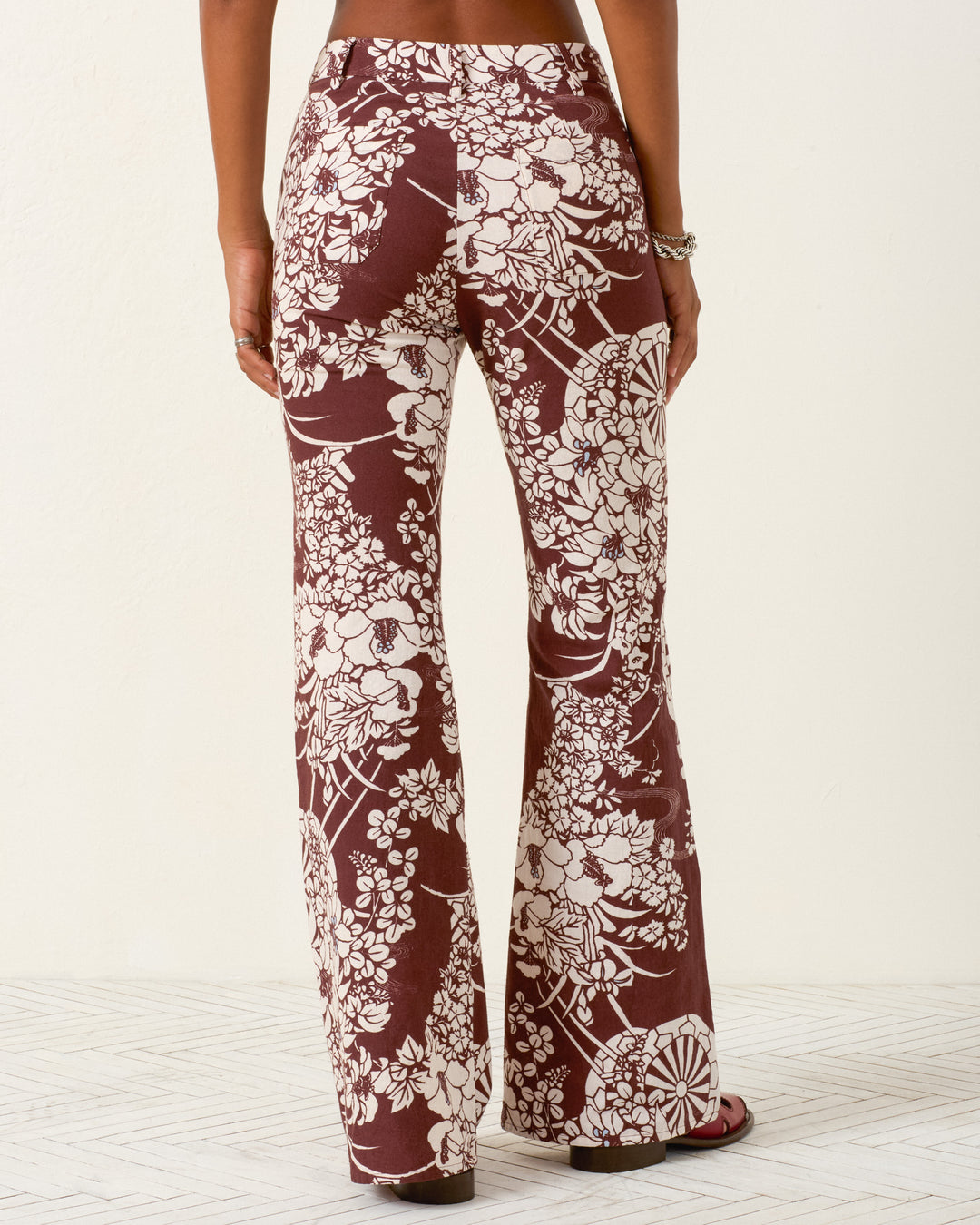 Person wearing the Bunny Auburn Spoke Jean—a high-waisted, brown flared pant with white hanaguruma floral print—stands with their back to the camera against a light background.