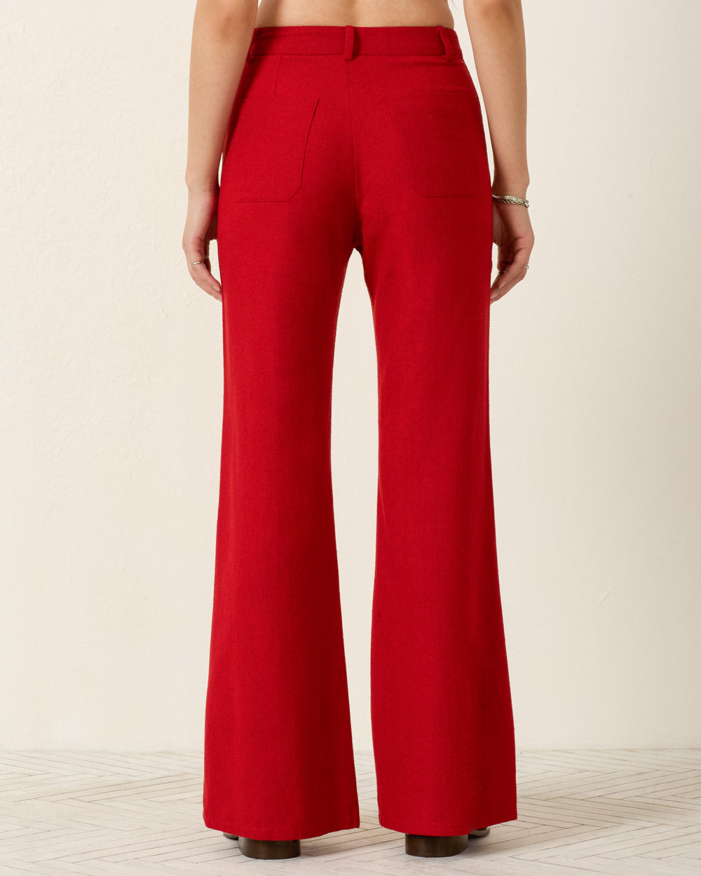 A person faces away, wearing Bunny Russet Silk Jean—high-waisted, red pants with a slim flared leg and back pockets—paired with black shoes against a neutral background.