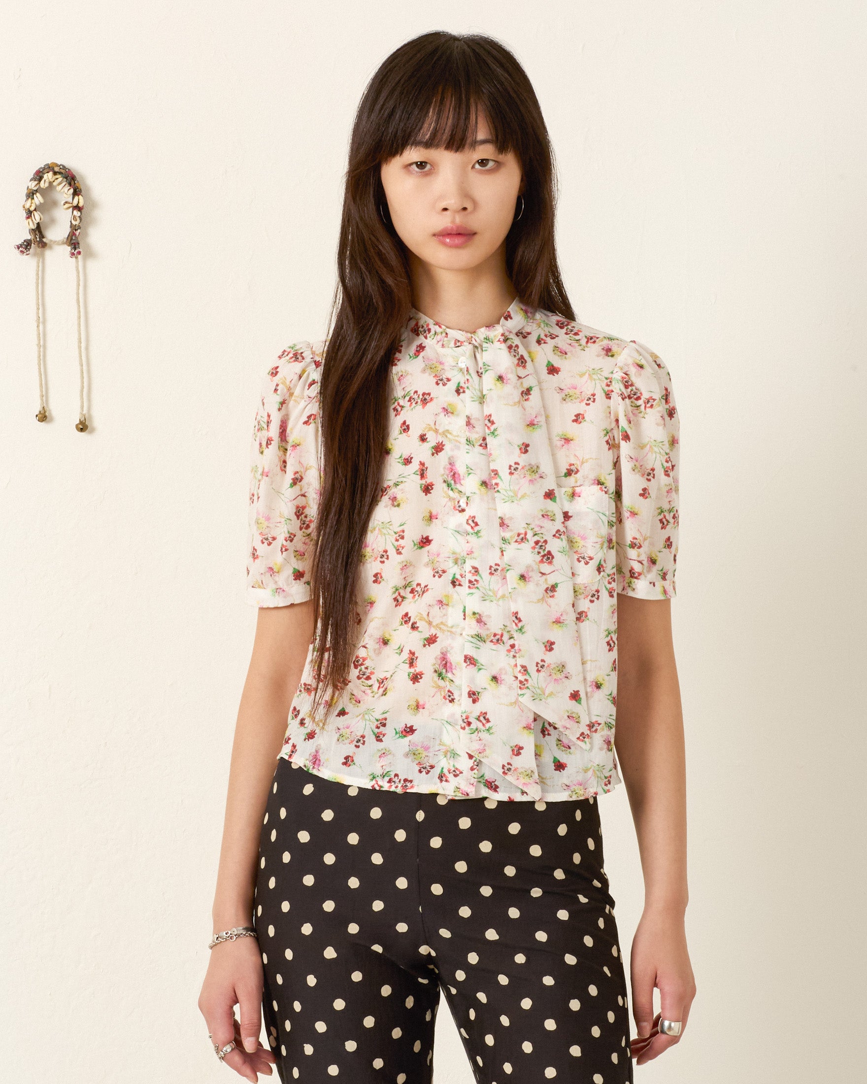 A woman stands before a plain wall, wearing the Chance Wild Garden Shirt—a silky modal blouse with a floral motif—paired with black and white polka dot pants. A small wall hanging appears to her left.