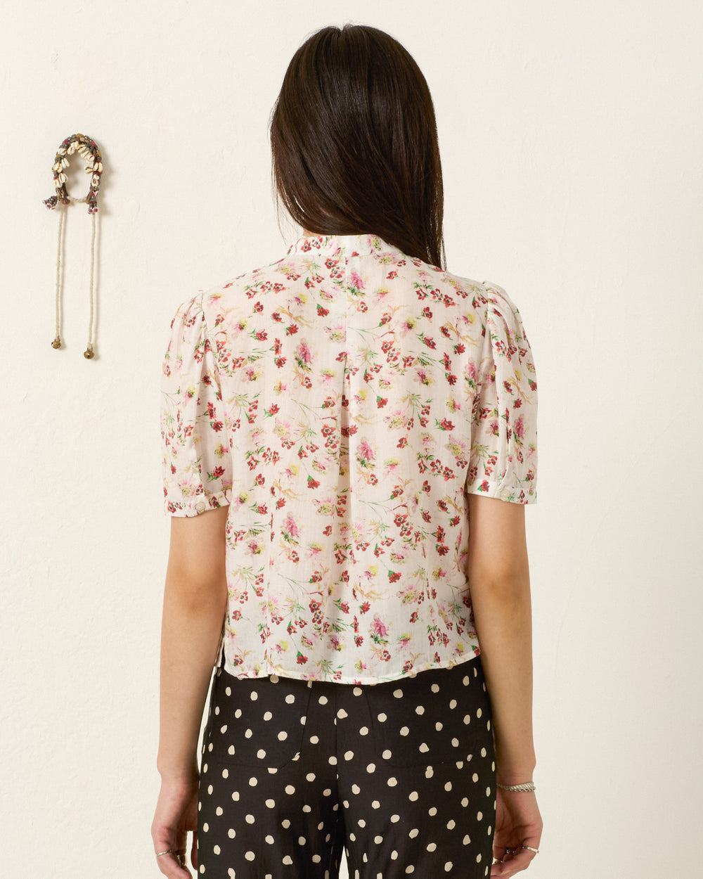 A woman faces away, wearing the Chance Wild Garden Shirt—a silky modal blouse with a floral motif—paired with black pants with white polka dots; a decorative wall hanging appears on the left.