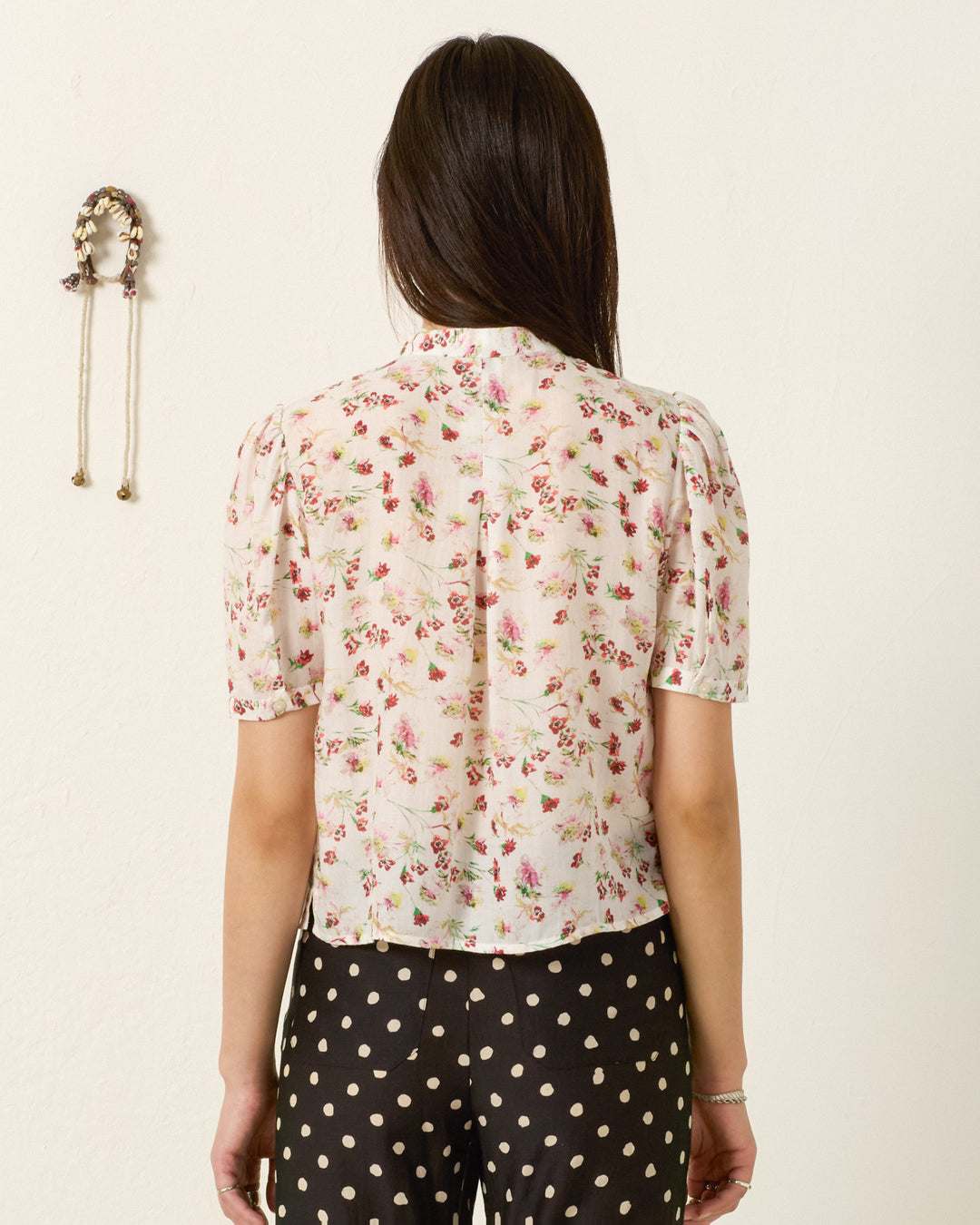 A woman faces away, wearing the Chance Wild Garden Shirt—a silky modal blouse with a floral motif—paired with black pants with white polka dots; a decorative wall hanging appears on the left.