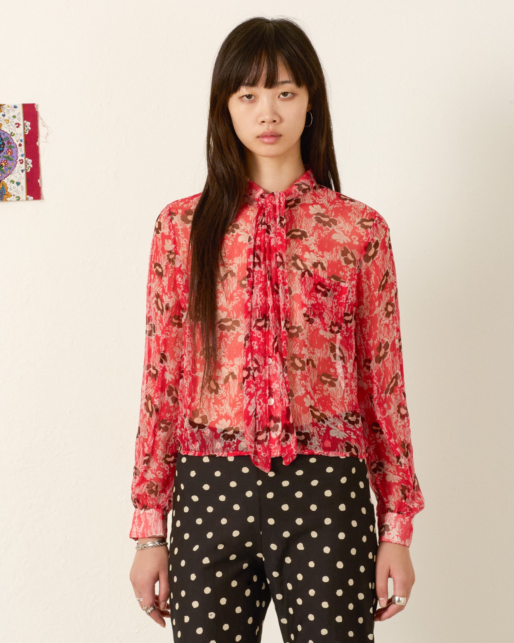 Indoors, a person wears the Chancery Rougewood Shirt—a semi-sheer red blouse with a '70s floral motif and pussy-bow tie—paired with black pants featuring white polka dots, and looks directly at the camera.