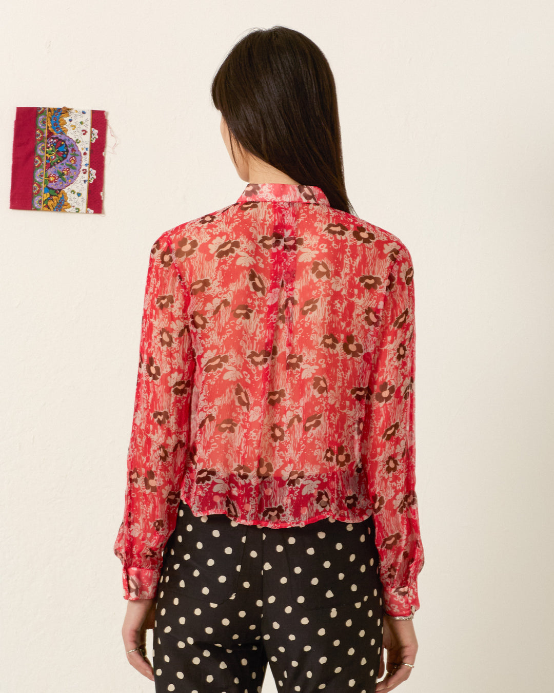 A person stands facing away, wearing the Chancery Rougewood Shirt—a semi-sheer red blouse with a '70s floral motif—and black pants with white polka dots. A small decorative fabric is pinned to the plain wall on the left.