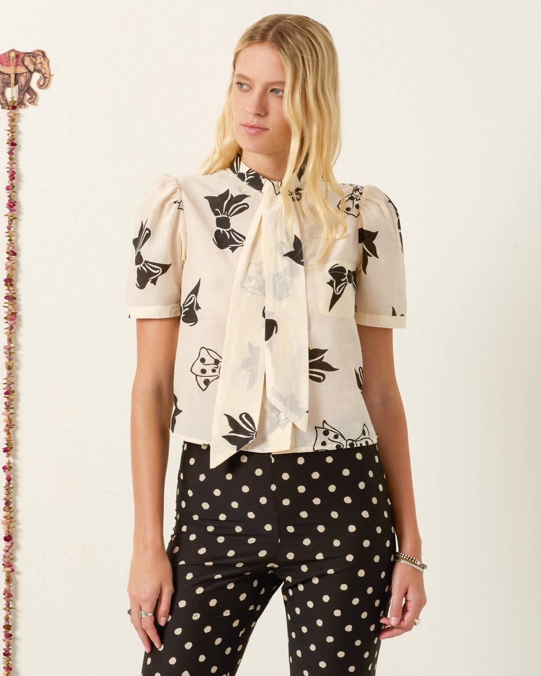 A woman models the Chance Black Tie Shirt—a short-sleeved, hand-block-printed blouse with floral design and bow detail—paired with black polka dot pants, standing indoors against a plain wall.